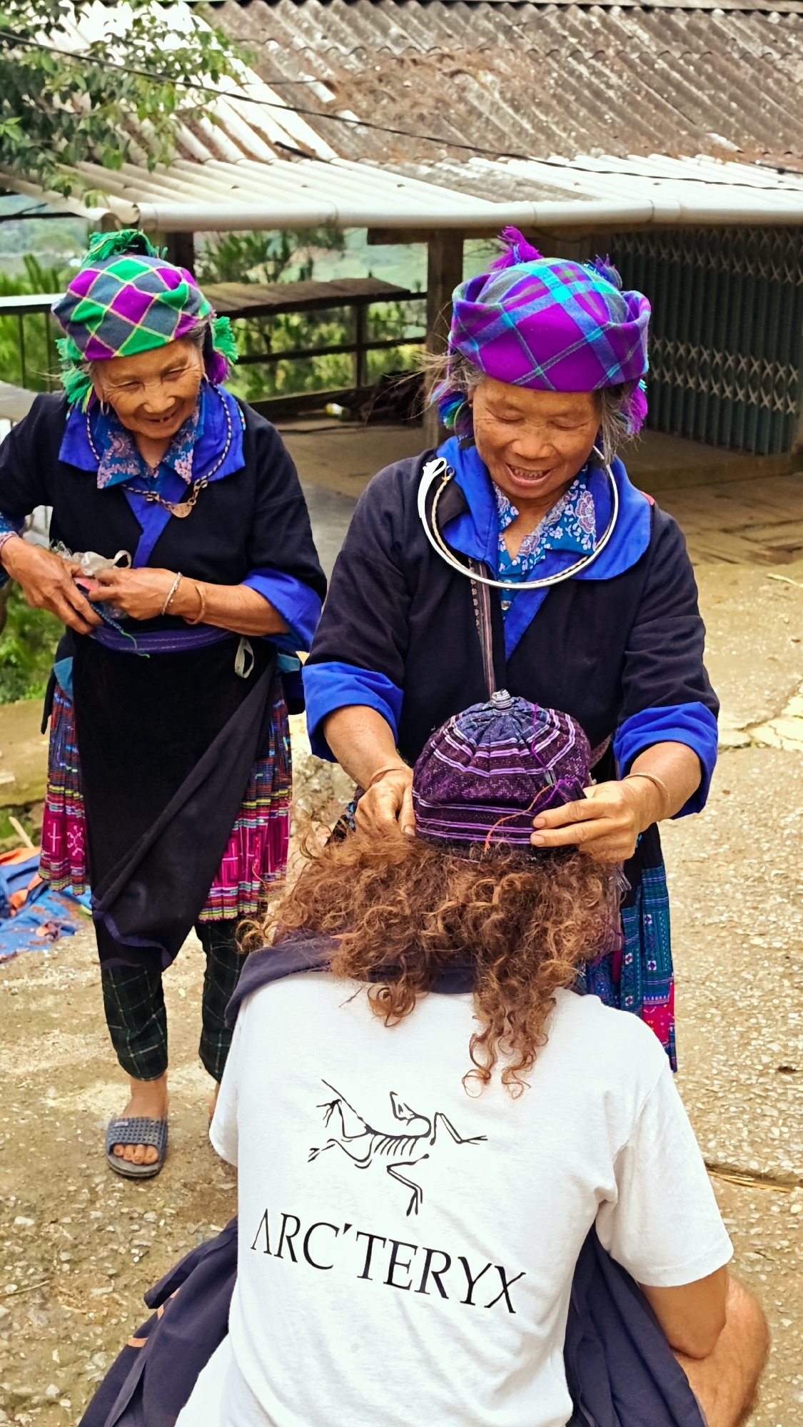 The image captures three individuals in an outdoor setting in Dế Xu Phình, Vietnam.
In the foreground, a person with their back to the camera, possessing long, curly, reddish-brown hair, is seen from the shoulders down. This individual wears a white short-sleeved t-shirt with a black "ARC'TERYX" logo and a skeletal bird graphic printed on the back. A dark blue fabric is draped over their shoulders.
Two elderly women, dressed in traditional attire, are positioned in the mid-ground. The woman on the left wears a black long-sleeved top with blue trim, a blue patterned scarf, and a vibrant green and purple checkered headscarf with green tassels. She has a gold-colored pendant visible on her chest and a gold-colored bracelet on her left wrist. Her lower attire consists of a dark skirt with multi-colored horizontal patterns and patterned green trousers, ending in gray sandals on her feet. She is smiling and her hands are positioned near her waist.
The woman on the right, also smiling, wears a similar black long-sleeved top with blue trim and a blue patterned scarf, topped with a purple and blue checkered headscarf with purple tassels. A large, ornate silver neck ring extends from her neck down her chest. She has gold-colored bracelets on both wrists. Her skirt is dark with blue and purple patterned trim. This woman is leaning forward, using both hands to place or adjust a small, dark purple, intricately patterned cap on the head of the person in the foreground.
The ground is an uneven, light brown dirt surface. In the background, there are two visible building roofs, one light gray and corrugated, the other darker. Green foliage is present on the left side of the image, and a dark, possibly metallic, grate or barrier is visible on the far right. Wooden structures are also discernible in the mid-background. The scene is illuminated by natural daylight.