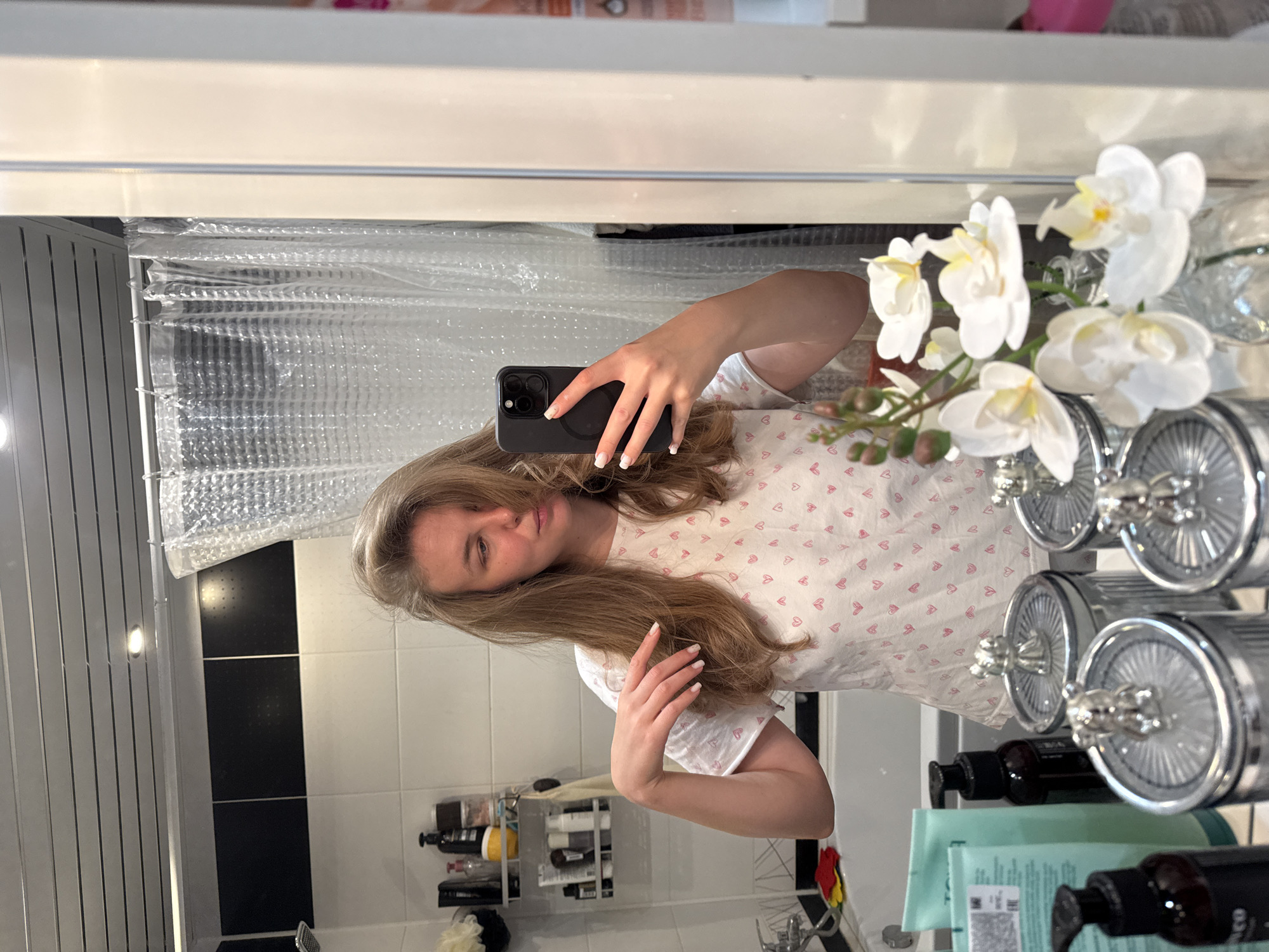 A young woman with long, light brown hair takes a mirror selfie in a bathroom. She is wearing a white t-shirt with a pattern of small pink hearts. Her left hand is holding a black smartphone, and her right hand is resting on her arm. In the background, a clear shower curtain with a textured pattern is visible. To the right of the woman, a decorative branch with white orchids is placed in a clear vase. The bathroom appears to have black and white tiled walls. On a shelf in the background, several bottles of toiletries are arranged. The lighting suggests it is daytime. The overall mood is casual and relaxed.