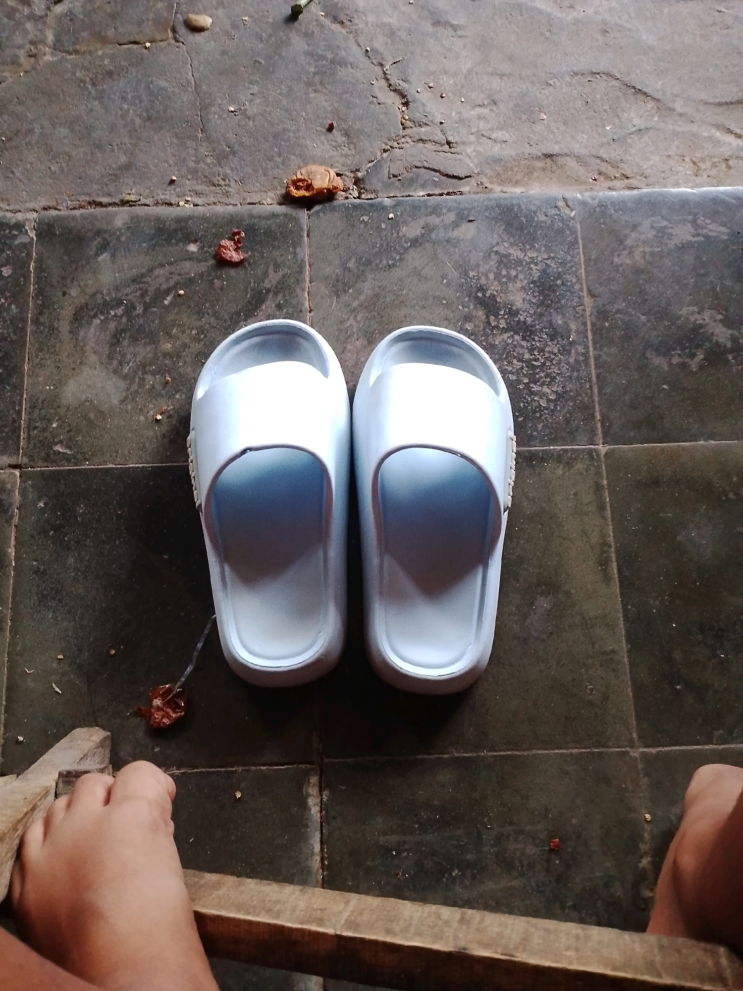 In Gebang, Indonesia, a pair of light blue slide sandals are placed on a tiled floor. The floor is dark and appears worn, with visible grout lines separating the tiles. Scattered on the floor are small pieces of debris, including what looks like dried leaves and some reddish-brown specks. To the left and right of the frame, parts of a person's feet are visible, suggesting they are either wearing the sandals or about to put them on. A wooden plank is positioned in the foreground, partially obscuring the view of the feet. The overall scene is domestic and casual, possibly an outdoor or semi-outdoor area.