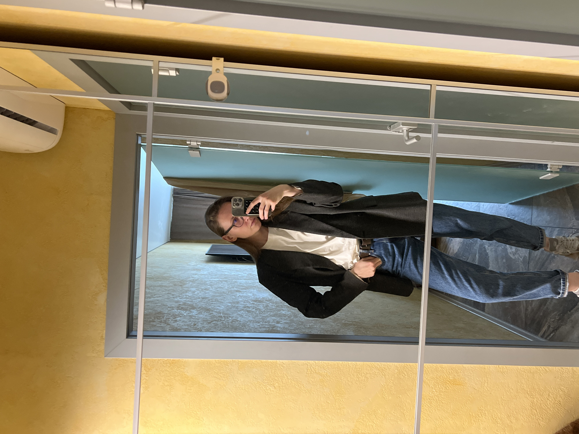 A young person with glasses, a white t-shirt, a dark blazer, and blue jeans is reflected in a set of mirrors in a room with textured yellow walls. There is a white air conditioning unit mounted high on the left wall. The room visible in the mirrors has a dark gray floor. The location is in Перепечино, Russia.
