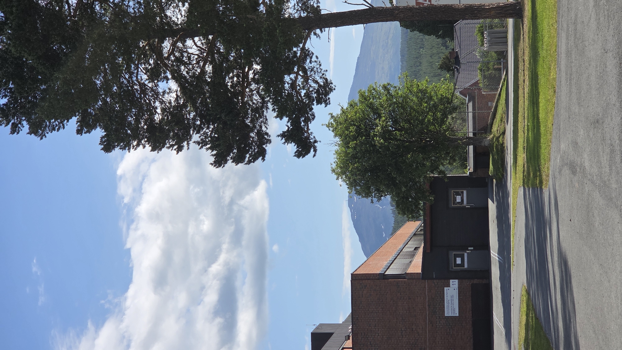 This image depicts the exterior of a brick and dark-colored building with a paved walkway and grassy areas in the foreground. A tall, mature pine tree stands prominently on the right side of the frame, with its branches extending into the blue sky dotted with white clouds. In the background, a lush, green forest covers rolling hills, and a distant mountain range with patches of snow is visible under the bright daylight.
The building appears to be a modern facility, possibly a school or administrative complex, with several dark-paneled doors and windows. A sign with text, partially obscured, is attached to the brick section of the building. The overall setting suggests a serene, semi-rural environment.
The scene can be identified as the area surrounding Nord-Østerdal videregående skole in Tynset, Norway. The architectural style of the building, the type of coniferous trees, the presence of the snow-capped mountain in the background, and the general landscape are consistent with the geographical and environmental characteristics of Tynset. The presence of a paved area typical of a school or public institution further supports this identification.