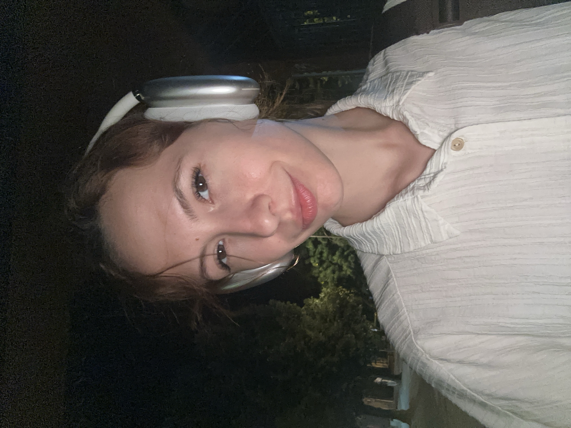 A woman with light skin and brown hair wears silver headphones and a textured white button-down shirt. She is positioned outside in a dark environment, with greenery visible in the background. The image orientation is sideways. The location appears to be Novi Sad, Serbia based on the plant and building styles, although specific landmarks are not visible.
