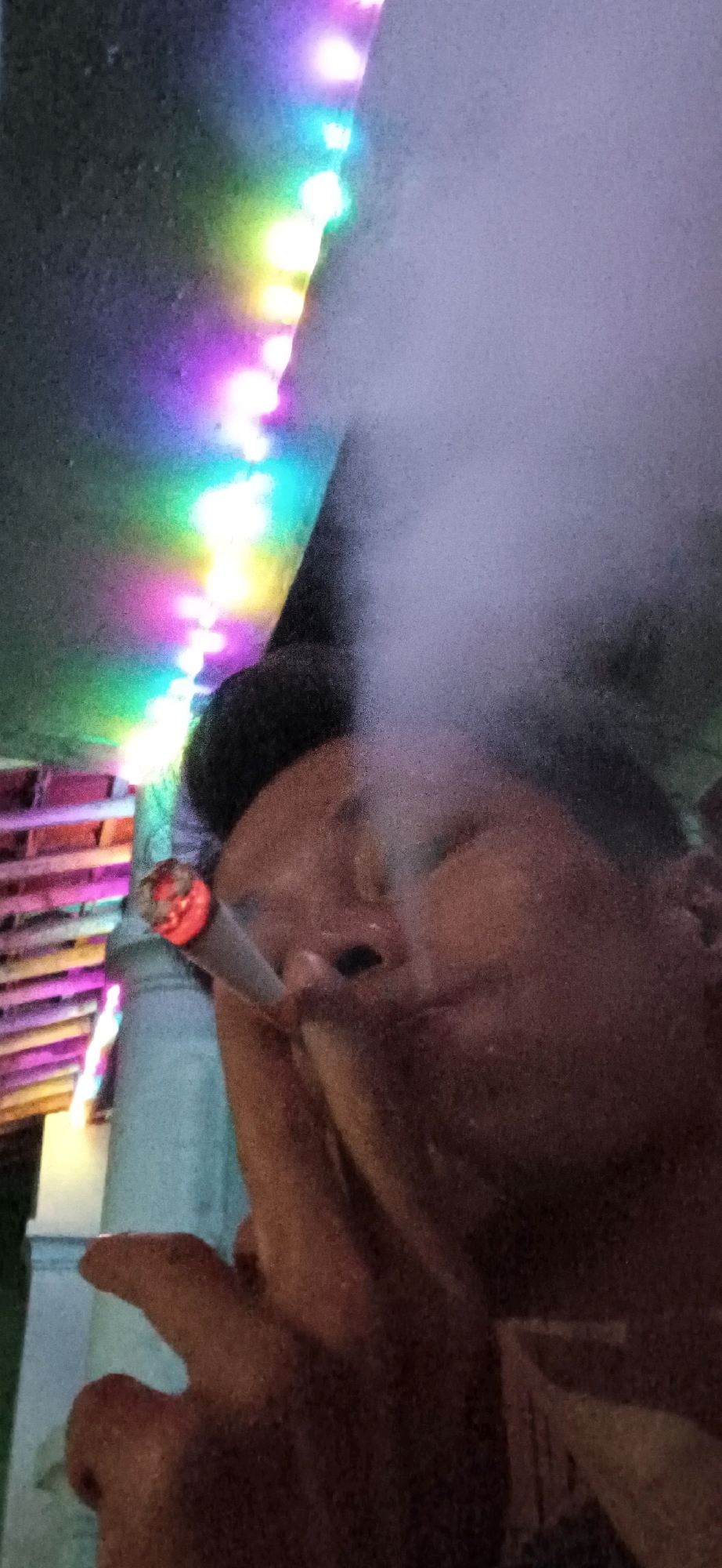 The image captures a close-up of a person smoking, with a prominent plume of smoke obscuring a portion of their face. The subject is holding a cigarette to their lips, and the tip of the cigarette is glowing red. In the background, a string of colorful lights, predominantly purple, green, and white, is visible against a dark surface. To the left, a portion of a structure with horizontal planks, possibly made of wood and painted in various shades of pink and yellow, is also apparent. The overall lighting is dim, with the colorful lights creating an atmospheric glow. The setting appears to be outdoors, possibly in a public space or backyard in Wonokerso, Indonesia, indicated by the ambient lighting and the visible architectural elements.