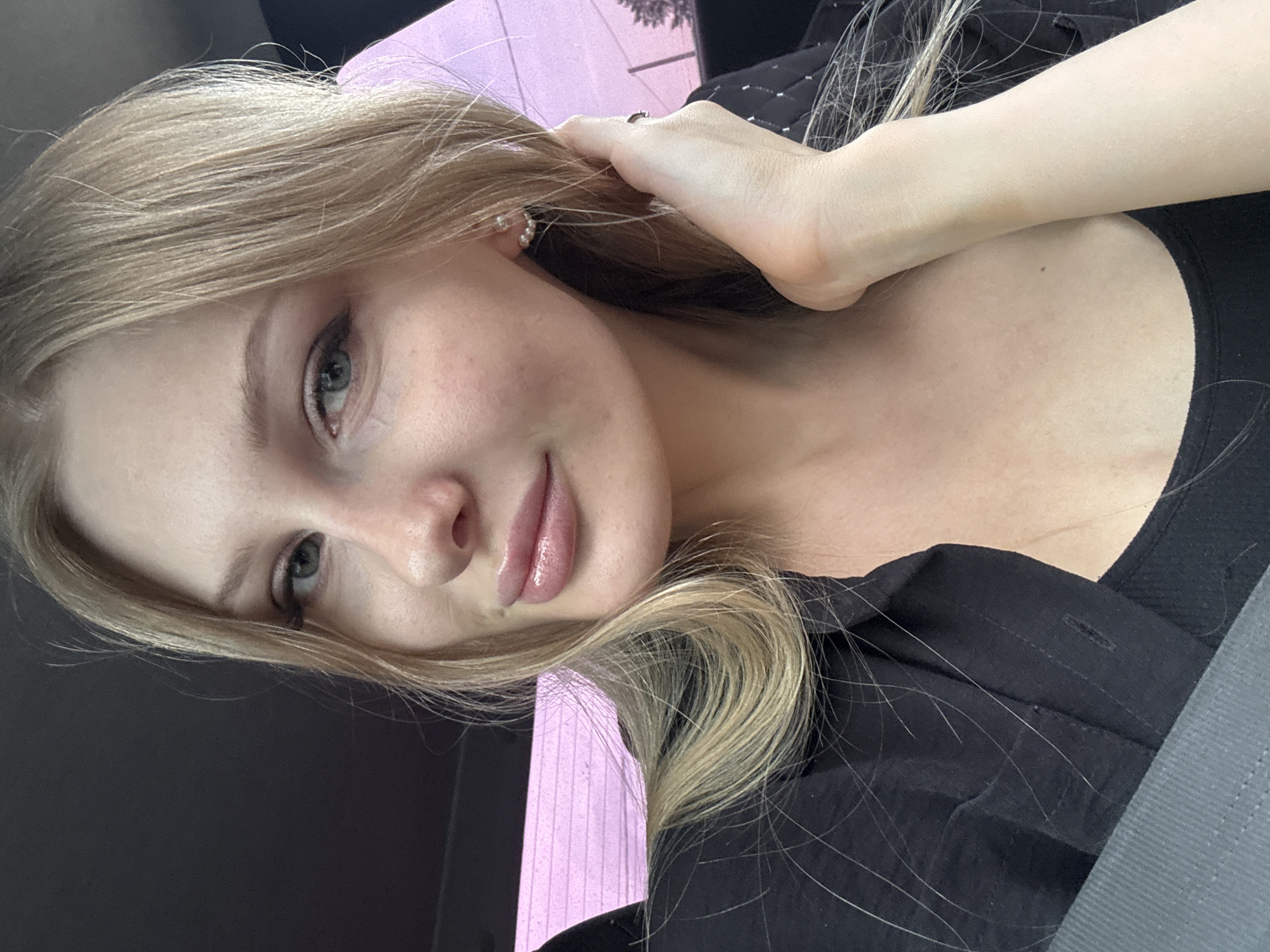A close-up, angled headshot captures a light-skinned young woman with blonde hair and light-colored eyes. She wears a black collared shirt. Her left hand is raised, touching her hair near her left ear, which has a small earring. The background includes a mix of pink-ish light and darker areas, potentially indicating an interior space. The exact location is not visually discernible. It is difficult to tell from the image whether the subject is located in Perm, Russia.