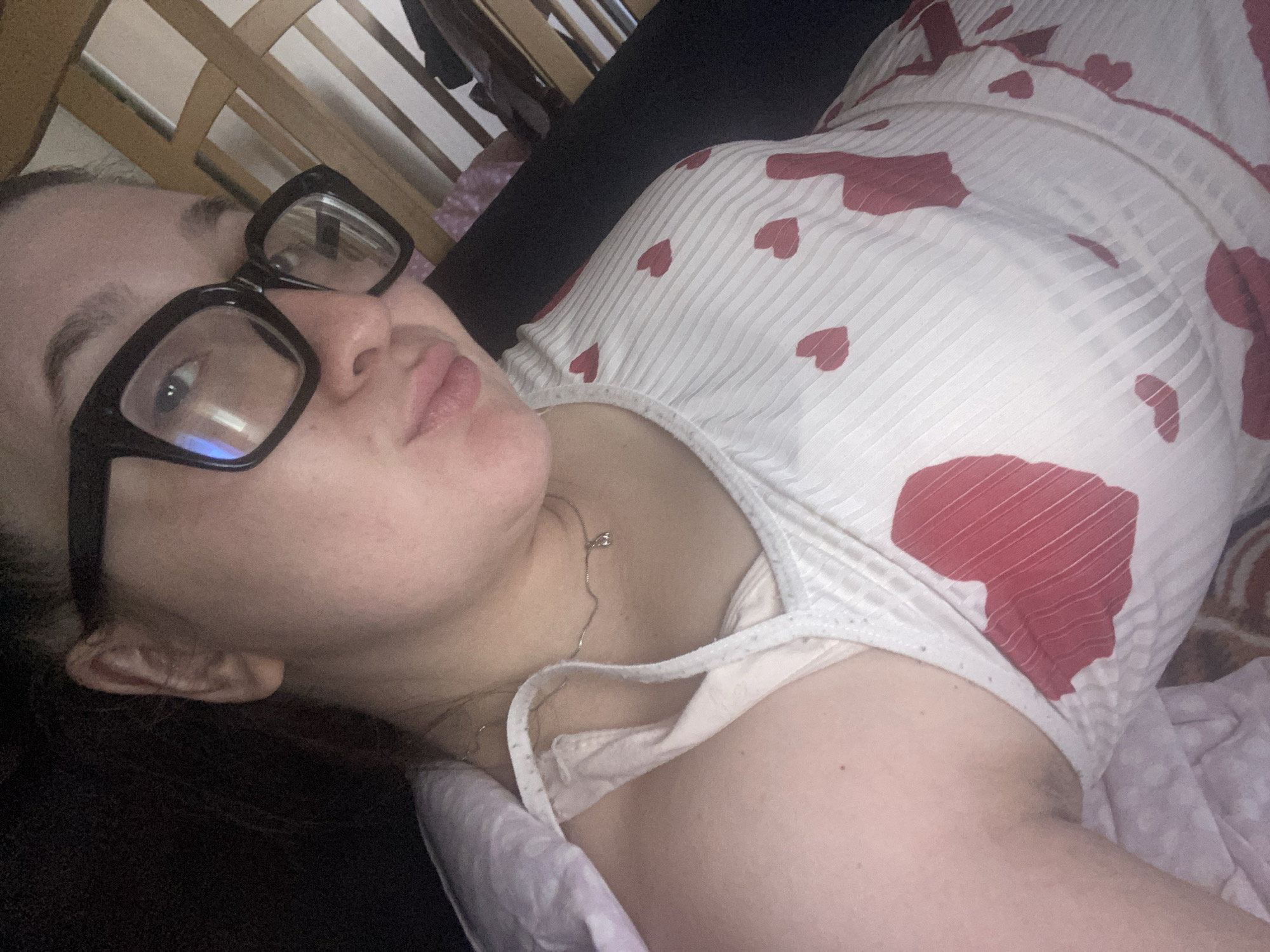 A woman with fair skin, wearing black-framed glasses, is lying on a bed. She is wearing a white top with red heart patterns. A thin silver necklace is visible around her neck. Part of her bra strap is also visible. The bed has a black headboard with wooden vertical slats. The bedding includes a lavender pillow and a patterned blanket. The location is not specified.