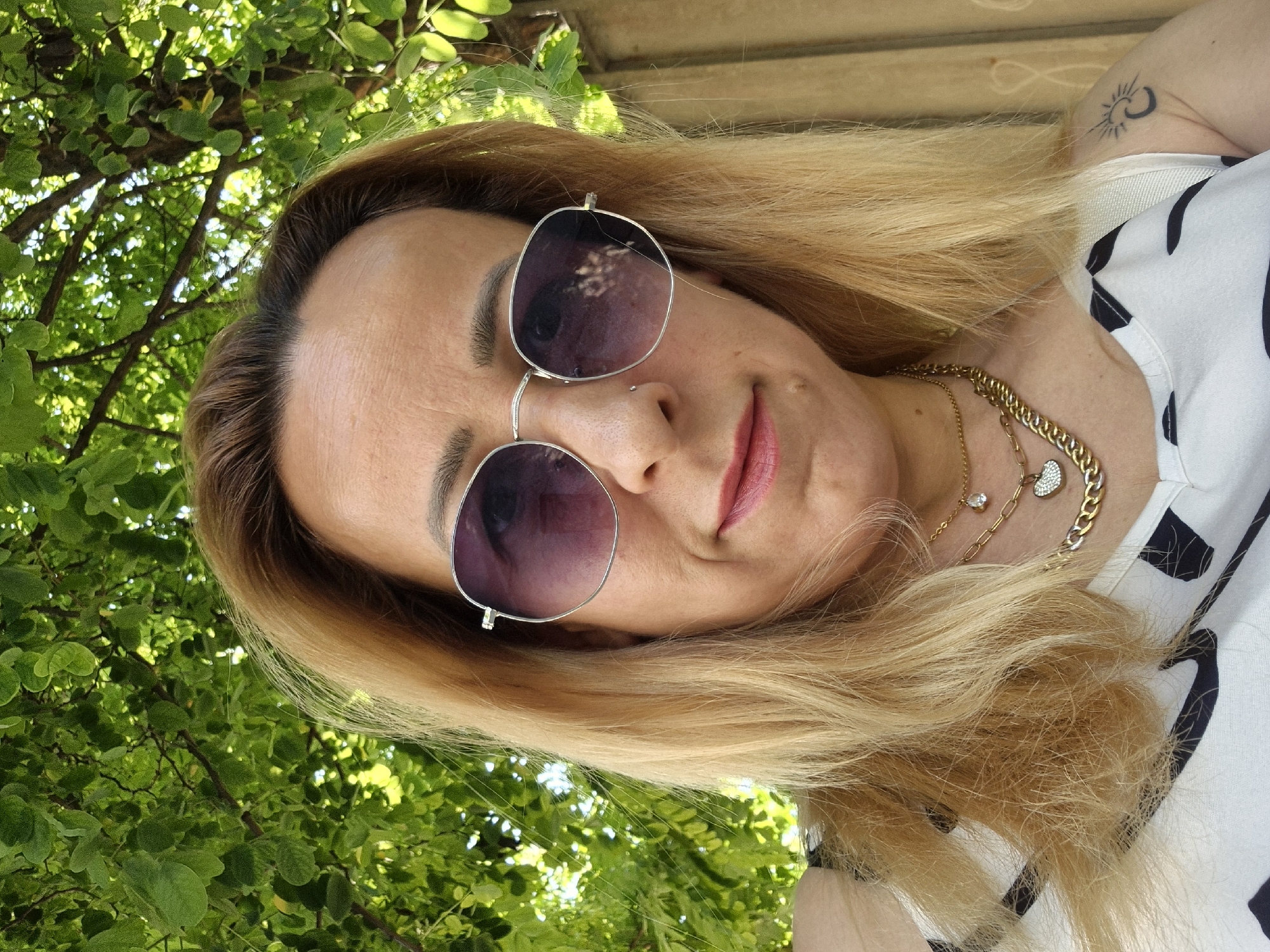 A medium shot captures a woman with blonde hair and sunglasses looking at the camera. She is outdoors, with lush green foliage visible in the background. The woman is wearing a white and black patterned top and several gold necklaces. A tattoo of a crescent moon and sun is visible on her left shoulder. The lighting suggests it is daytime, with sunlight filtering through the leaves.