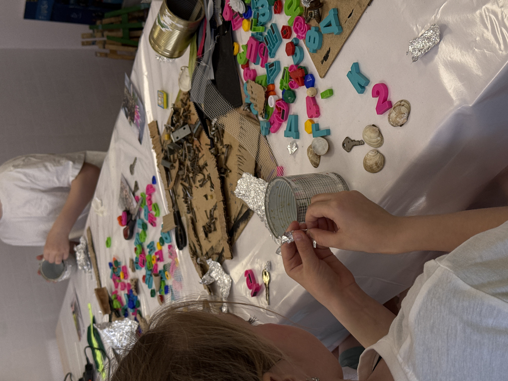 The image depicts a close-up view of a craft activity in progress, likely in a home setting. Two individuals are present, at least partially visible. One person, wearing a white t-shirt, is seen from the chest up and has their hair in the frame. This person appears to be manipulating tin foil and adhering it to a tin can. The second person, also in a white t-shirt, is holding a tin can, their arms visible. A white tablecloth covers a table, which is scattered with craft supplies including colorful plastic letters and shapes, shells, keys, bits of cardboard, mesh, and other metallic and plastic items. There are several tin cans, some wrapped in tin foil. The location is not discernable from the image, but has been requested to be placed in Chaikovsky, Russia.