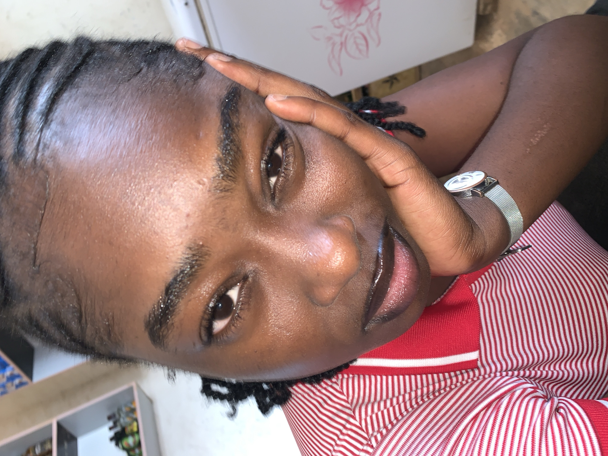 A close-up, slightly tilted portrait features a young person, likely a woman, with dark skin and braided black hair. She looks directly into the camera with a calm and composed expression. Her lips are slightly parted, revealing a glossy finish.
Her left hand is raised, with fingers resting gently against her forehead and temple. On her left wrist, she wears a silver-toned watch featuring a mesh band and a white circular face with dark markings. She is dressed in a red and white striped top, possibly a polo shirt. Her braided hair is styled neatly, with some baby hairs visible along her hairline. The skin on her forehead and face appears shiny or hydrated.
The background is softly out of focus, suggesting an indoor or sheltered environment. Vague shapes of light-colored surfaces and indistinct objects, potentially shelves, are visible. One detail captured in a crop shows a faint, light-colored drawing resembling floral or leafy patterns on a white background surface. Another cropped detail highlights a section of her braided hair secured with a red and black string. A faint scar is also visible on her right forearm in one of the close-up shots.
The image appears to be a posed photograph or selfie, taken during what seems to be daytime hours. The general location is New Stadium, Nigeria. No discernible text is visible within the image.