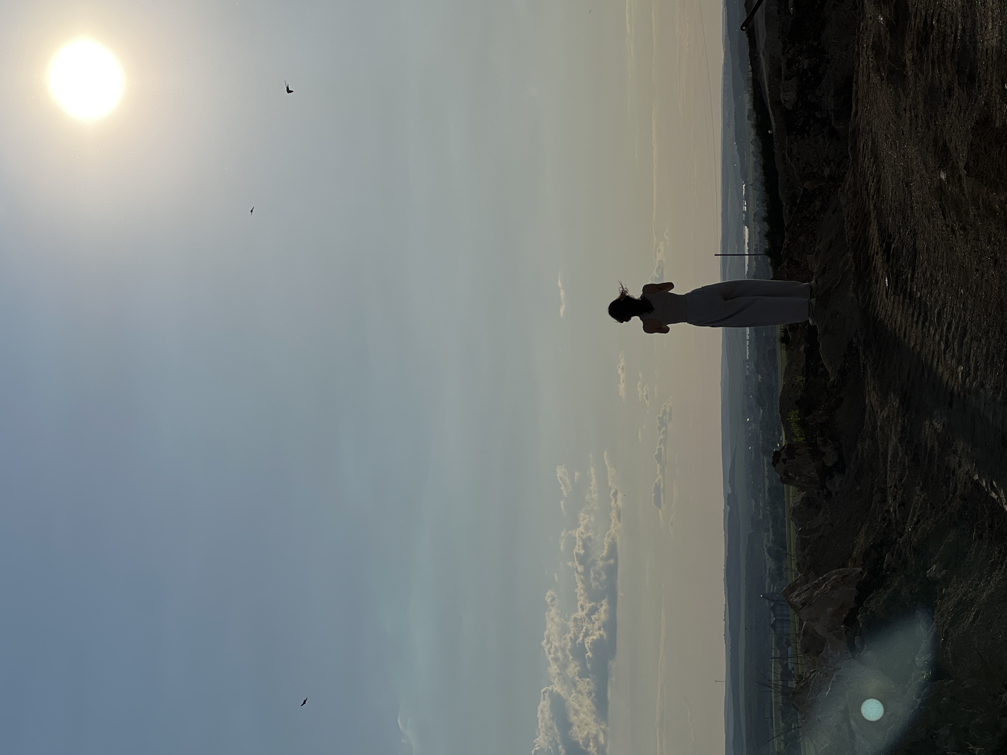 A lone figure stands silhouetted on a mound of earth, facing away from the camera towards a vast landscape under a bright, hazy sun. The person, wearing light-colored loose-fitting pants and a tank top, has dark hair that appears to be windblown. The ground in the foreground is rough and dark, possibly disturbed earth or a construction site, with large rocks scattered around. In the mid-ground, a rolling landscape stretches out, dotted with trees, fields, and some distant buildings. The sky is a pale blue with wispy clouds and several birds in flight. The sun, high in the sky and slightly off-center, casts a warm, diffuse light over the scene. The overall impression is one of solitude and contemplation against a wide-open natural backdrop. The location is identified as Khomivka, Ukraine.