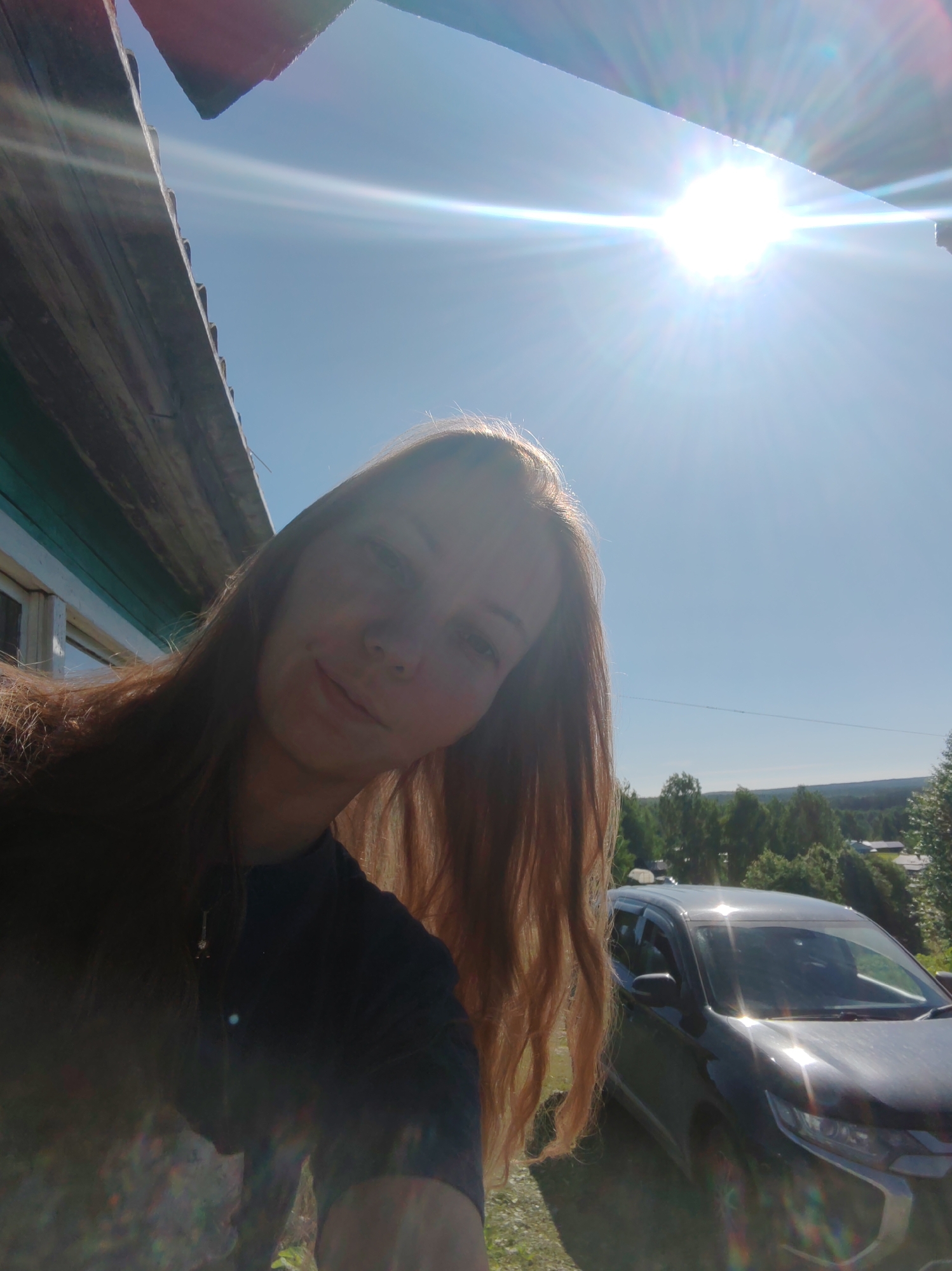 An outdoor, back-lit selfie depicts a young woman with long, reddish-brown hair smiling subtly towards the camera. She wears a dark top with a visible pendant and is positioned to the left of the frame. Strong sunlight originates from the upper right, causing significant lens flare and obscuring parts of the woman's face and the environment. To the upper left, a weathered wooden eave or roofline of a building, partially painted teal, is visible. The sky above is clear blue. In the middle-ground to the right, a dark SUV, possibly a Mitsubishi Outlander, is parked on what appears to be a dirt or gravel surface, reflecting sunlight. The background features a distant landscape of dense green trees, indicative of a forest or wooded hills, with scattered white and light-colored structures (houses) nestled among them. A faint power line is visible across the distant horizon. The scene is located in Чухлэм, Russia.