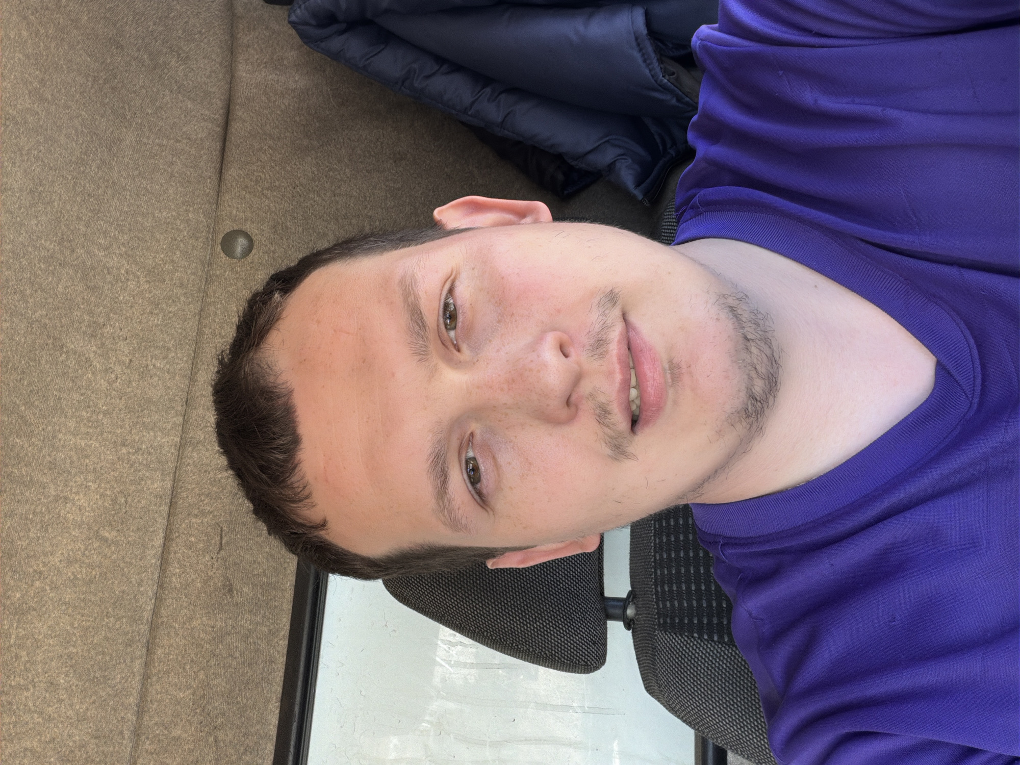 A young adult male is featured in a close-up portrait, likely a selfie, looking directly at the camera with a neutral to slightly smiling expression. He has short, dark hair, light skin with visible freckles, and light brown eyes. He wears a plain, long-sleeved purple shirt with a crew neck and has light stubble on his chin and upper lip.
The background suggests an indoor setting or the interior of a vehicle. To the man's right, there is a textured, light brown or tan wall/paneling with a small, circular metallic fixture. To his left, a dark grey mesh-fabric headrest or seat back is visible. A dark blue padded jacket is draped in the upper right background. The lower part of the image shows a blurred, light-colored surface, possibly a window or a reflective surface. The lighting indicates it is daytime. There are no specific visual cues within the image itself to confirm the location as Chaikovsky, Russia. No text is visible in the image.