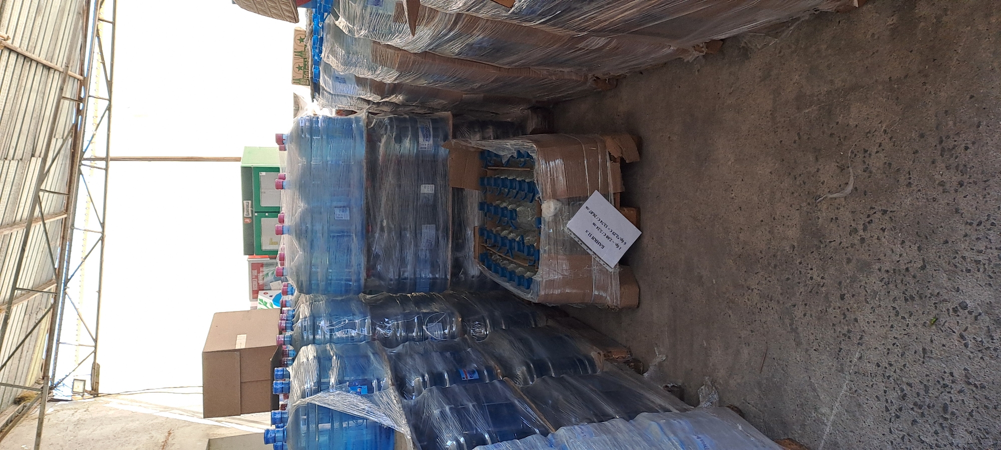 The image depicts a storage area with stacks of bottled water. The bottles are large, clear plastic containers with blue liquid, capped with pink. They are shrink-wrapped onto pallets, forming tall stacks. In the foreground, a pallet of smaller water bottles with blue caps is visible. A white paper label with text in Bulgarian is affixed to one of the boxes of small bottles. In the background, there are large cardboard boxes and green and white structures that appear to be display units or kiosks. The environment suggests an indoor storage facility, possibly a warehouse or distribution center, with a concrete floor and a metal roof structure overhead. The scene appears to be located in Velika, Bulgaria, based on the provided context. There are no people visible in the image.