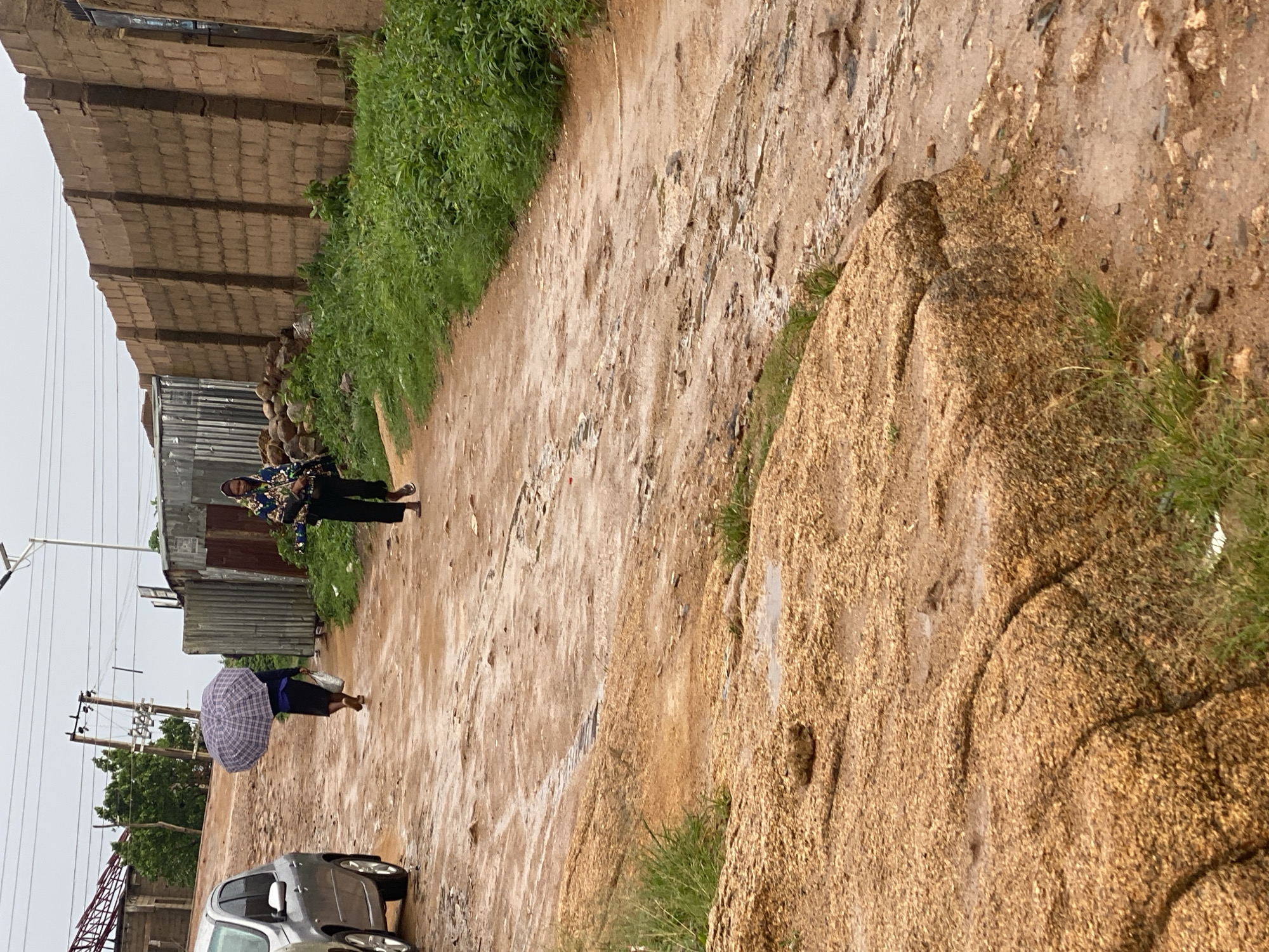 The image depicts a dusty, unpaved road in the city of Jos, Nigeria. The road appears wet, suggesting recent rainfall. In the foreground, a large, rough, tan-colored rock formation dominates the lower left of the frame. The road stretches into the distance, curving to the right.
Two individuals are visible on the road. The first, further up the road and to the left, is walking away from the camera, holding an open, checkered umbrella. They are wearing dark clothing and appear to be barefoot. To the right and closer to the camera, a second person is walking towards the camera, also appearing to be barefoot. This individual is wearing a dark-colored headscarf and a jacket with a colorful, patterned design, along with dark pants.
To the left of the road, a silver car is partially visible. The background features a low, rough brick wall on the right side, with some green vegetation growing along its base. On the left, there are corrugated metal structures and some trees. Overhead, power lines and a street light are visible against a cloudy sky. The overall impression is of a simple, somewhat neglected urban environment in Jos.