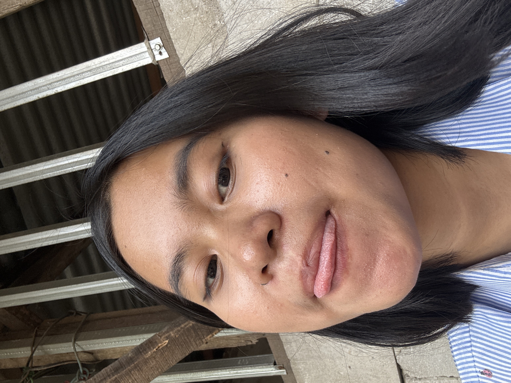 A close-up selfie shows a young East Asian woman with dark hair and dark eyes. She is wearing a blue and white striped shirt. Her tongue is sticking out slightly. The background appears to be outdoors, with a corrugated metal roof and wooden beams visible. The setting is consistent with an outdoor area in East Jakarta, Indonesia.