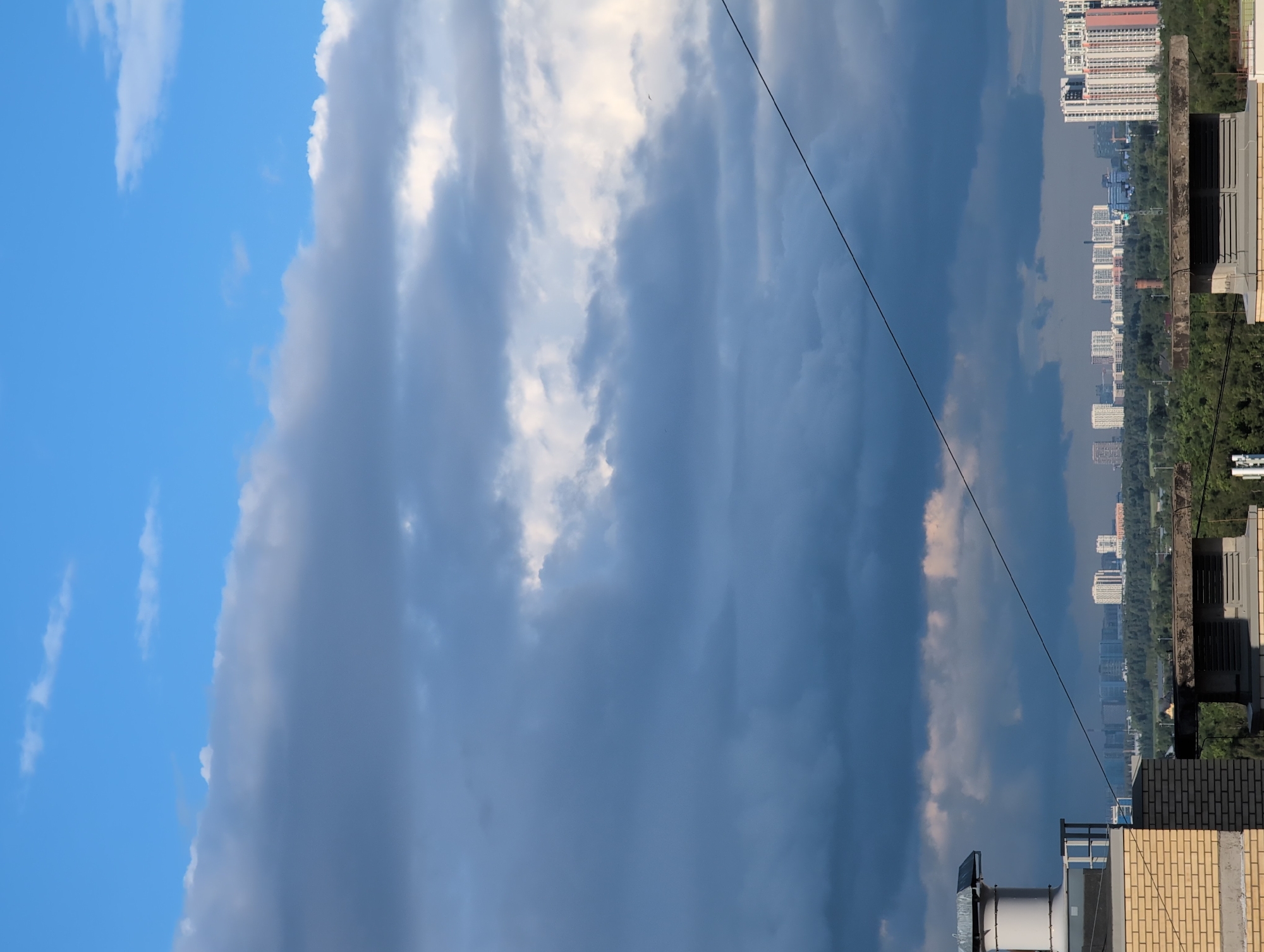 The image depicts a cityscape with a dramatic sky. The scene is oriented vertically, suggesting the photo was taken in portrait mode. The sky dominates the upper portion of the frame, transitioning from a bright, clear blue on the left to dark, heavy storm clouds on the right. Wispy white clouds are scattered throughout, catching the sunlight. A thin black wire stretches diagonally across the sky from the top right to the bottom left, a subtle but noticeable detail.
In the background, a dense urban landscape stretches horizontally. Numerous apartment buildings of varying heights and architectural styles are visible, interspersed with patches of green trees. The buildings suggest a relatively modern city. The foreground features the tops of two buildings, one with a brick facade and another with a light-colored, textured material. On the first building, there's a large, white industrial fan or ventilation unit. The second building has visible rooftop details like vents and metal structures.
The time of day appears to be late afternoon or early evening, as indicated by the contrast between the bright blue sky and the darkening storm clouds, and the quality of light. The weather seems to be transitioning from fair to stormy. There are no people visible in the frame. The overall emotion evoked is one of atmospheric drama and urban expanse. The location is hinted to be Saint Petersburg, Russia, by the urban architecture. No text is visible in the image.