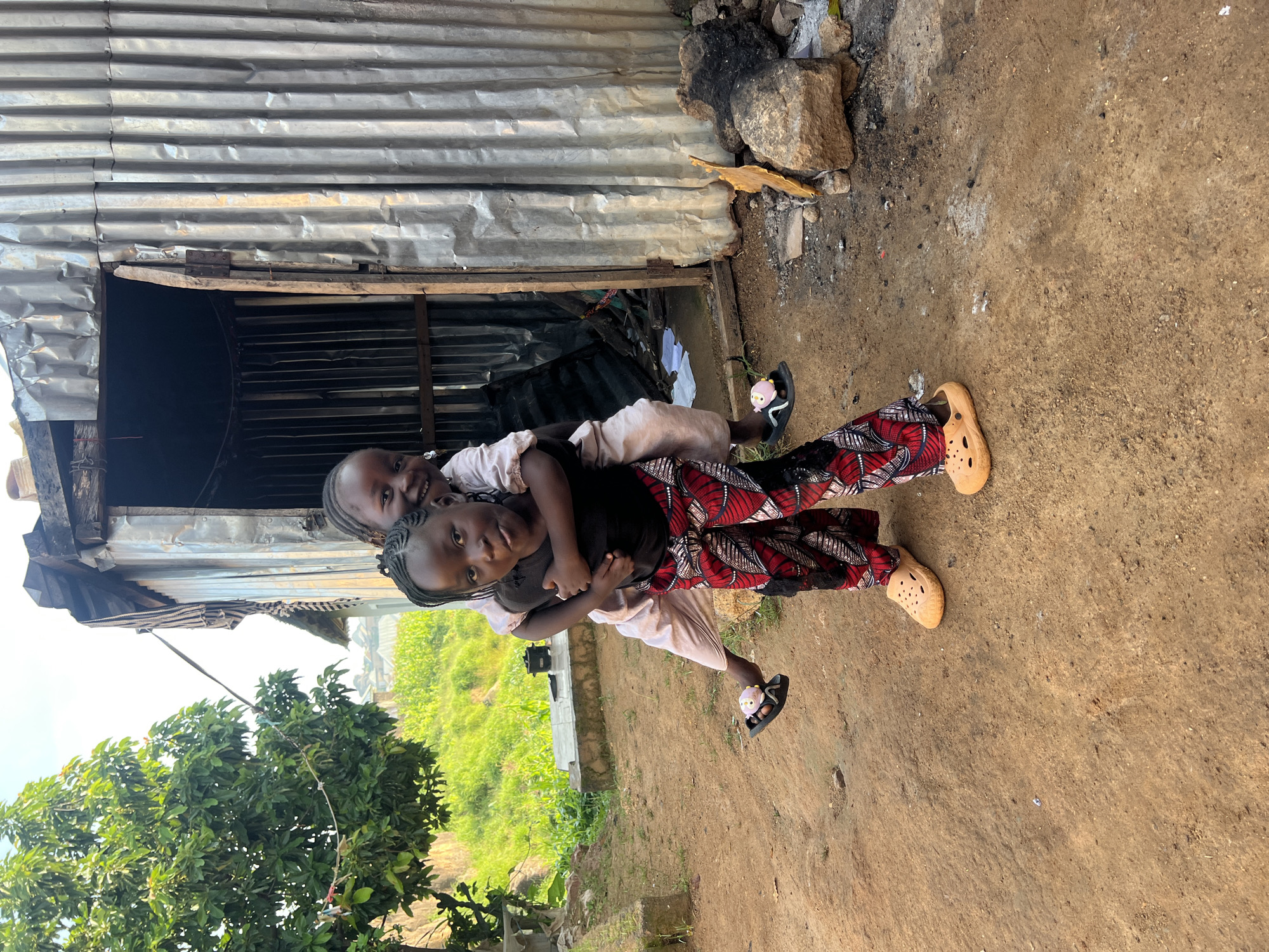 Two young girls are in the foreground outside a corrugated metal structure in Jos, Nigeria. The girl in front is facing the camera, giving a piggyback ride to the girl behind her, who is also facing the camera and smiling. Both girls are wearing colorful clothing and sandals. The structure behind them appears to be a makeshift building with a wooden door. The ground is dirt, and there are some rocks and debris in the background. A tree is visible to the left of the scene.