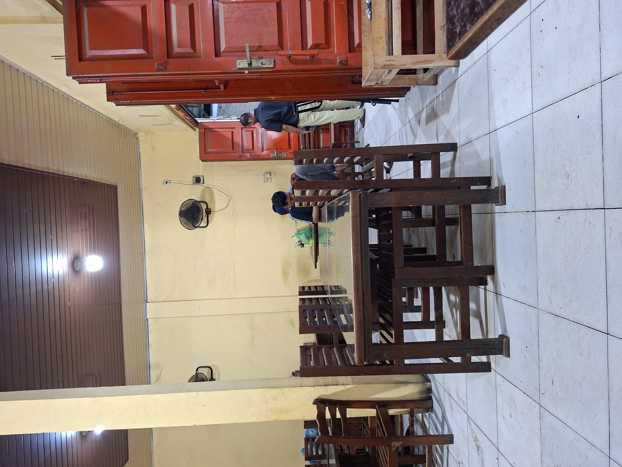 An indoor scene, possibly a casual eatery or waiting area in Keudegeudong, Indonesia, is depicted, with the image rotated 90 degrees clockwise. The setting features light yellow walls, a tiled floor with some visible wear, and a ceiling that combines brown wooden panels with light-colored sections.
Several dark wooden tables and chairs, of a simple and sturdy design, are arranged in the space. Most tables are empty, but one in the mid-ground has a clear glass surface. Two men are present. One man, wearing a blue shirt and light-colored trousers, stands near a prominent red wooden door, looking downwards, possibly at a phone in his hand. Another man, in a dark shirt and grey trousers, is seated at one of the wooden tables, looking towards the left side of the frame.
Artificial lighting illuminates the space, with bright ceiling lights visible, suggesting it could be evening or a dimly lit interior. Two wall-mounted oscillating fans are visible, one in the mid-ground and another further back, indicating a need for ventilation. Power outlets and visible wiring are present on the walls. The overall atmosphere appears functional and unadorned. No visible text is discernible in the image.