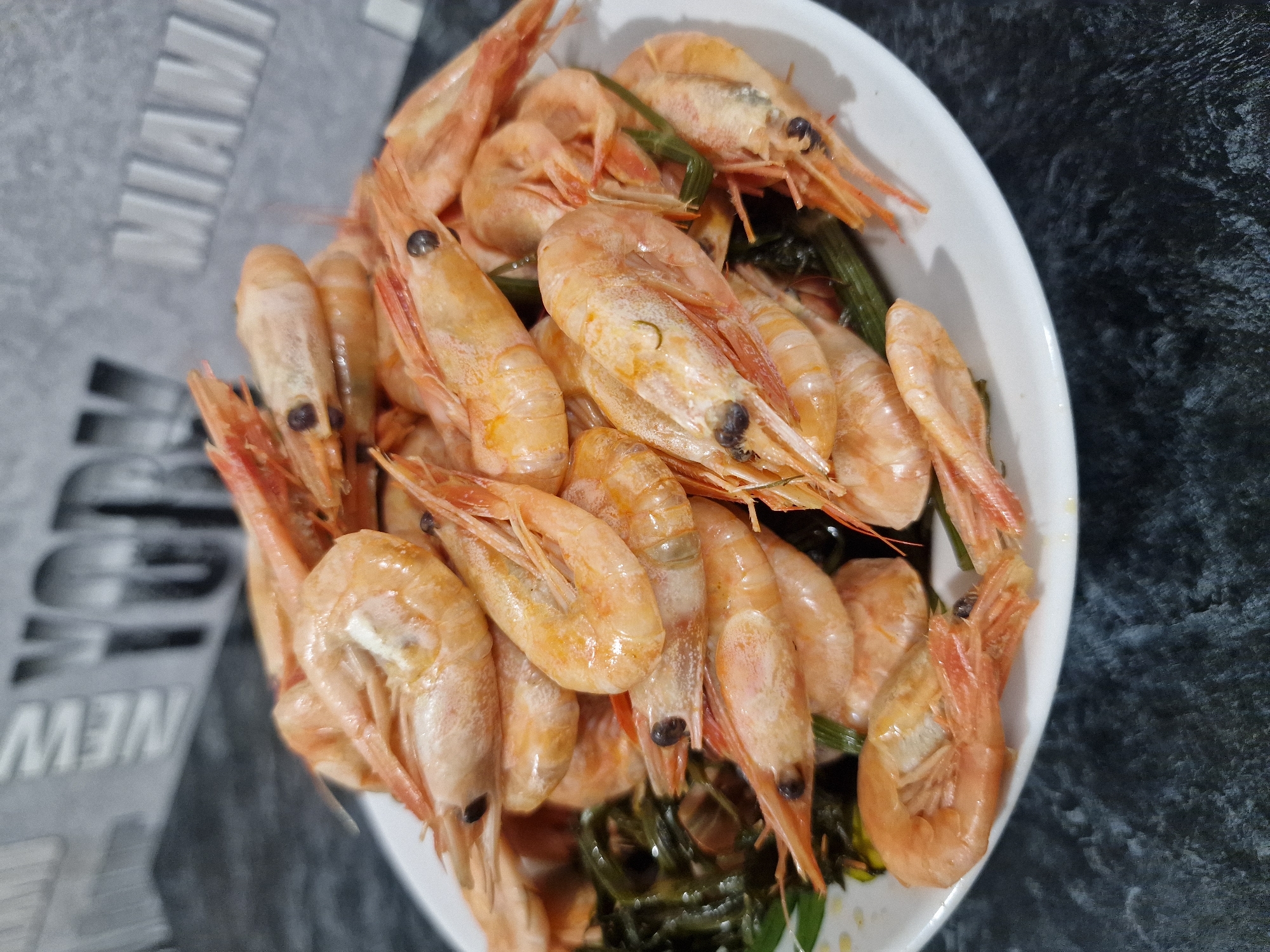 A white plate of cooked shrimp and green vegetables is placed on a dark speckled surface. The shrimp are pinkish-orange and piled on top of each other. The green vegetables appear to be seaweed or similar. In the blurred background, there is a wall with the words "NEW YORK" and "MIAMI" written in large, stylized letters. The overall lighting is dim. There are no people visible in the image. The location is not Kursavka, Russia; it is a generic indoor setting.