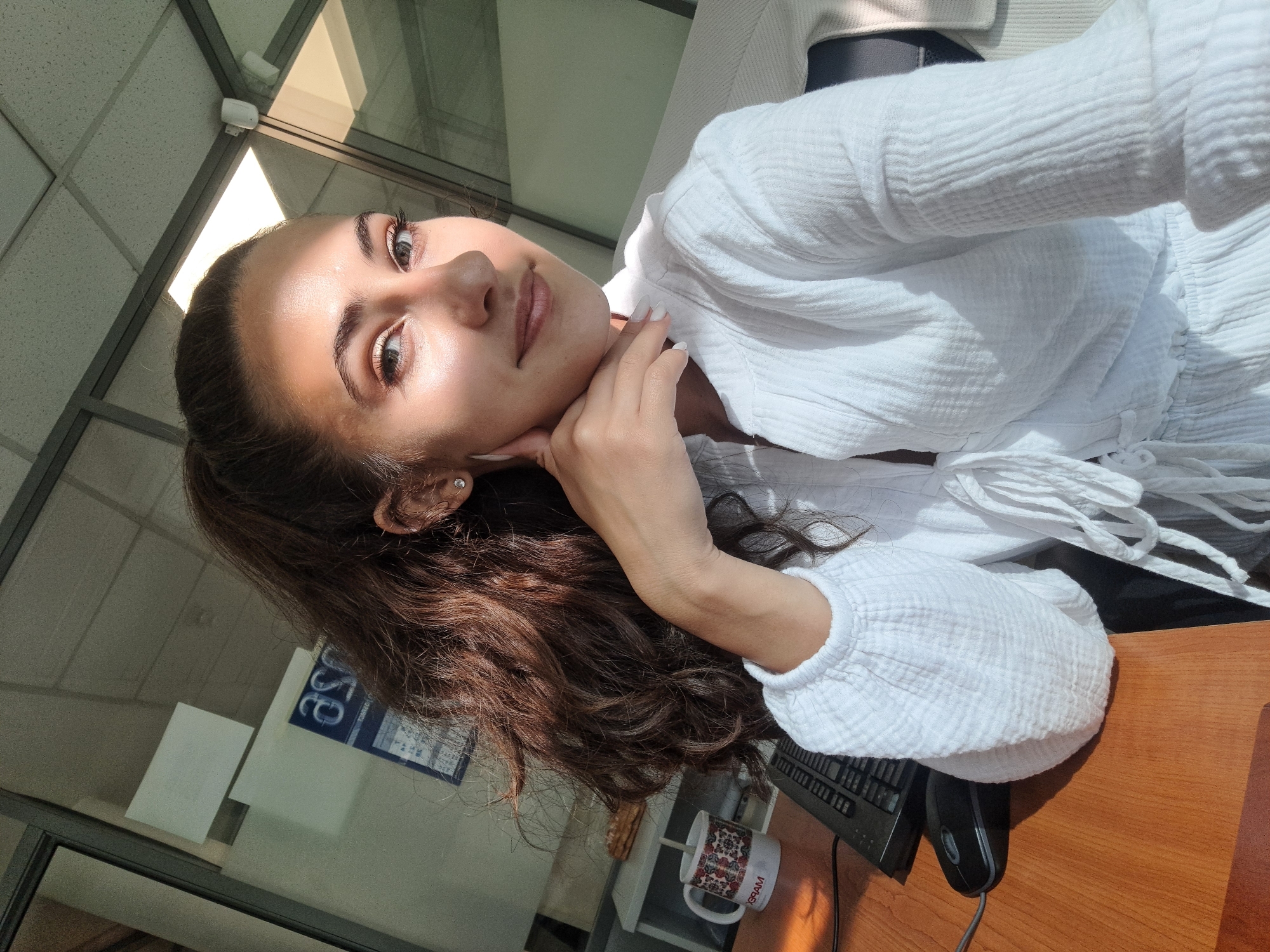 A young woman with long, wavy brown hair tied in a high ponytail is sitting at a wooden desk in an office in Sofia, Bulgaria. She is wearing a white, crinkled-fabric blouse with a tie closure at the front. Sunlight streams in from the right, casting a bright highlight across her face and upper body. She is looking towards the camera with a slight smile, resting her chin on her left hand. Her nails are painted white. On the desk in front of her is a computer keyboard, a black mouse, and a white mug with a colorful floral pattern and the text "MAPEX" visible. Behind her, a glass partition separates her workspace from another area of the office. A calendar with blue and white print is visible on the wall in the background. The office environment appears modern, with acoustic ceiling tiles and fluorescent lighting.