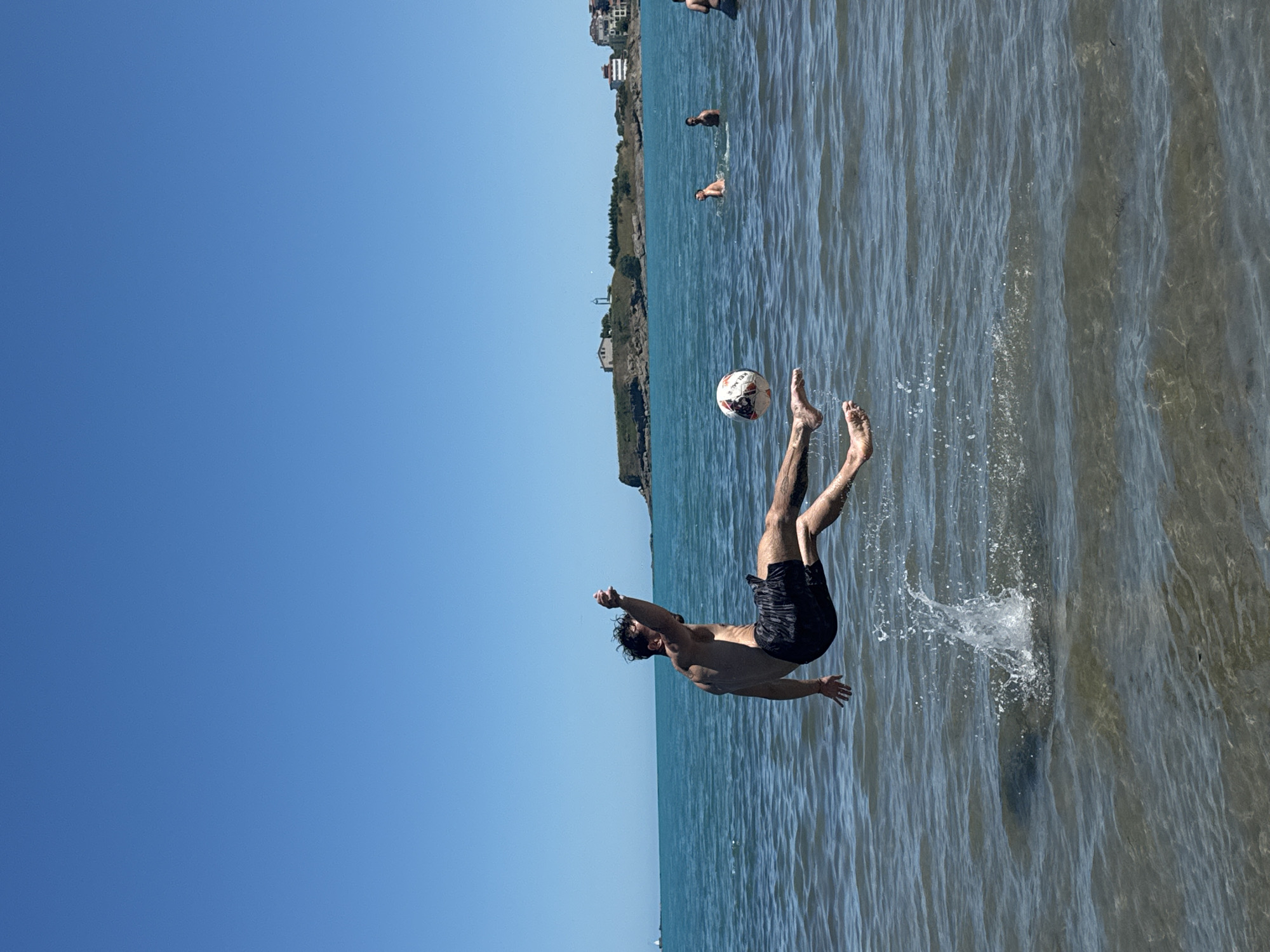 In the city of Tsarevo, Bulgaria, a young man is captured mid-air in the shallow waters of the sea, performing a skillful kick at a soccer ball. He is wearing black swim trunks, and his body is contorted as he propels himself upwards, creating a splash as his feet leave the water. The soccer ball, a white and black pattern, is airborne and positioned to be struck by his leg. The background reveals a clear blue sky and the tranquil blue sea, with a distant shoreline featuring buildings and a small hill. Several other individuals are visible in the water, appearing to be swimming or enjoying the beach. The scene is one of active recreation on a sunny day.
