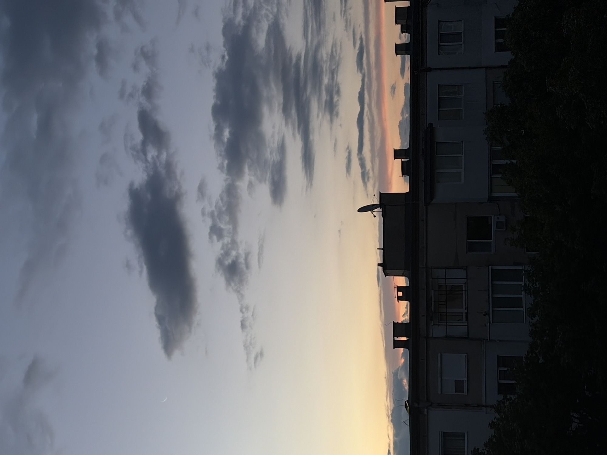 The image shows a twilight sky over a multi-story building. The sky displays a gradient of colors, transitioning from a pale blue at the top to warmer hues of orange and pink near the horizon. Several dark, wispy clouds are scattered across the sky, and a thin crescent moon is visible in the upper left quadrant.
The building's rooftop features multiple chimneys and a satellite dish on one of the upper structures. The facade of the building is visible below the rooftop, characterized by rows of windows, some of which are dark, suggesting they are unlit or viewed from the exterior. In the foreground, the dark silhouettes of trees obscure the lower portion of the building. The overall scene suggests an urban environment at dusk.