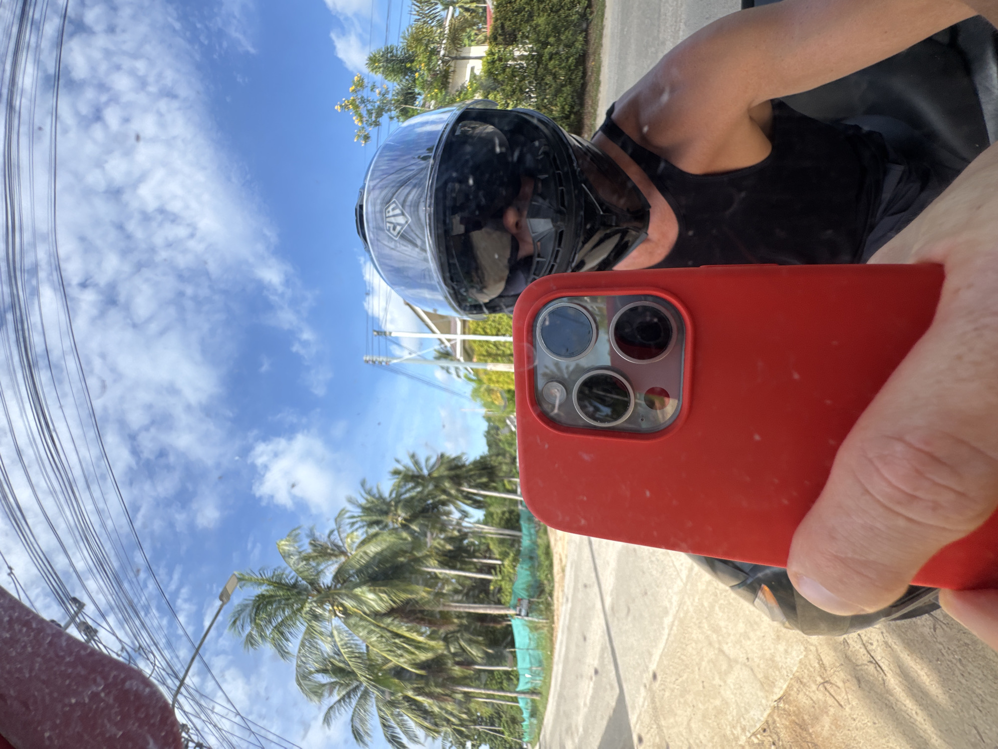 A selfie is taken by a person wearing a black motorcycle helmet and a black tank top. The person is holding a red smartphone up to take the photo, and the reflection of the person and the surroundings can be seen in the helmet's visor. The background shows a street lined with palm trees and buildings, under a blue sky with white clouds. The location is in Tambon Ko Pha Ngan, Thailand, evident from the tropical vegetation and architecture. The road appears to be paved, and there is a turquoise net fence along the side of the road. Power lines are visible above the trees.