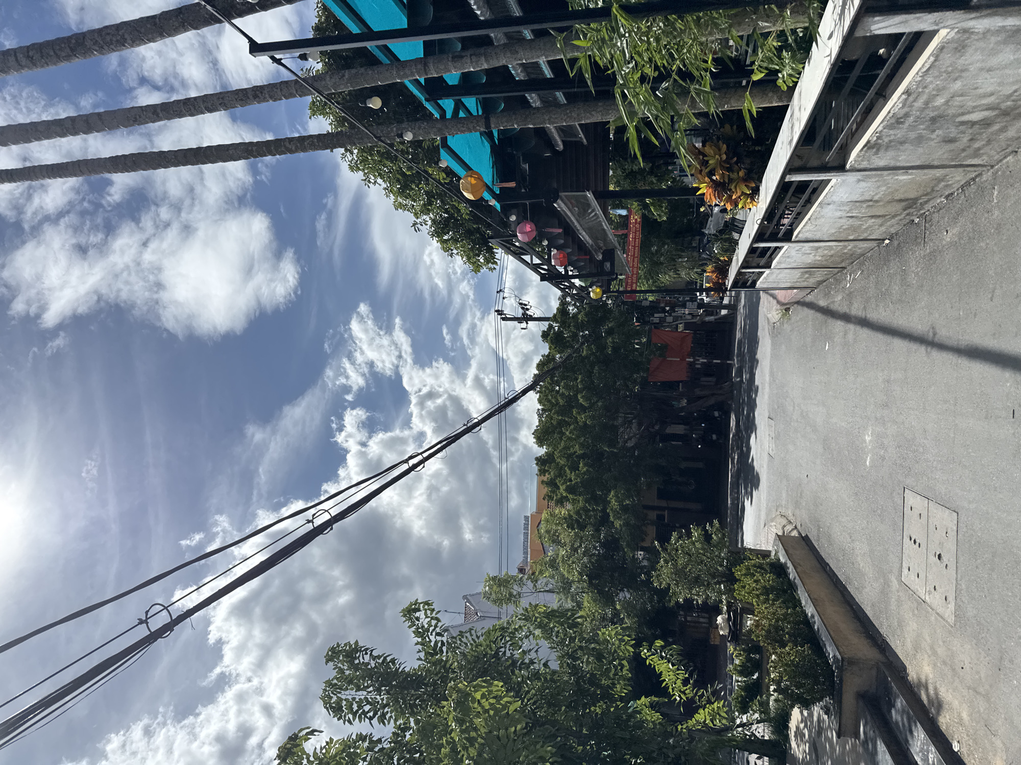 This image depicts a street scene in Hoi An, Vietnam, likely on a bright, sunny day with scattered clouds. The perspective is from a street-level view, looking forward down a paved road.
On the left side of the road, there are several lush green trees and a concrete planter box filled with manicured shrubs. Further back, a white building with a sloped roof is visible, and above it, an orange-tiled building with the text "A BOUTIQUE HOLIDAYS" can be seen. Overhead, power lines stretch across the frame, supported by utility poles.
The right side of the road features a textured concrete wall with a metal railing on top. Beyond the railing, several tall, slender trees with rough bark are prominent. A structure with a bright turquoise awning is visible, adorned with hanging paper lanterns in yellow, pink, and red. String lights with exposed bulbs are also present.
The road itself is asphalt, showing some wear. A manhole cover with a grid pattern is set into the pavement on the left. In the distance, the road curves slightly, with more trees and buildings lining the sides. The overall impression is a well-lit, somewhat tropical urban environment. No people are clearly visible in the frame.