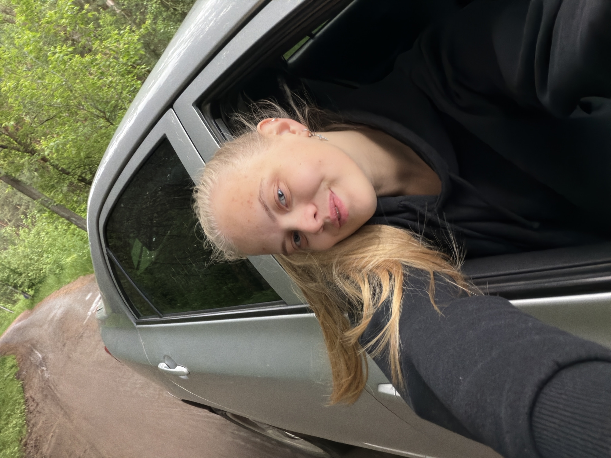A young person with light blonde hair and fair skin is leaning out of the open window of a silver-colored car. The individual is wearing a black hooded sweatshirt. The car appears to be on a dirt or gravel road surrounded by green vegetation, including trees. The location appears to be a rural or semi-rural environment, and there is no information available to indicate the scene's connection to the city of Chaikovsky, Russia. The road's surface suggests recent rain or damp conditions.
