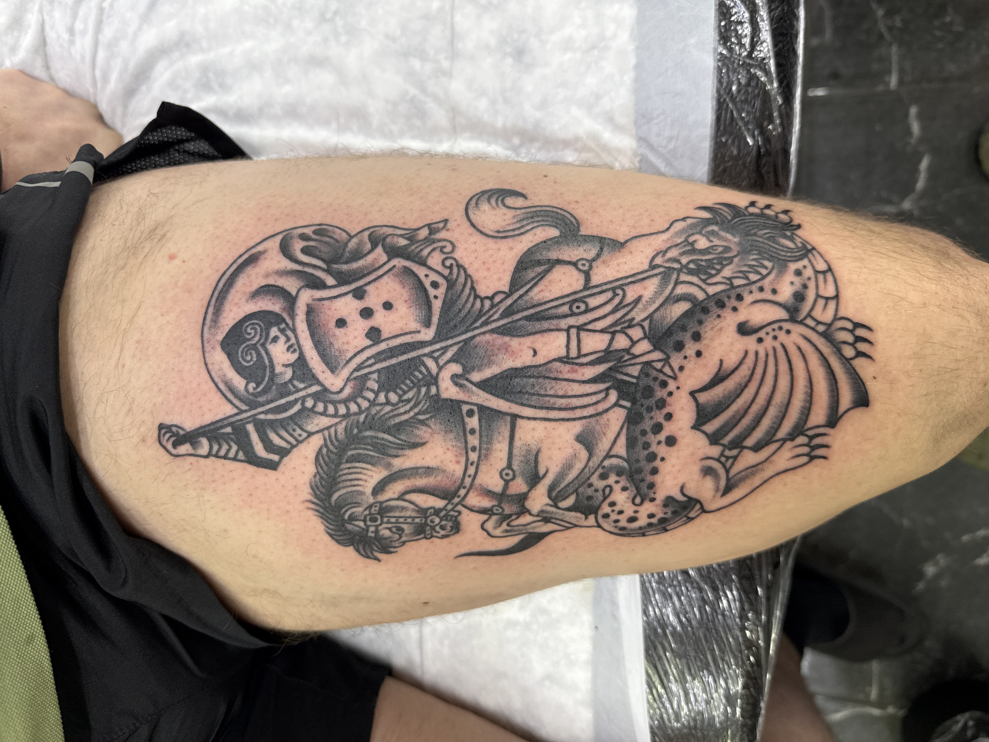 A black and grey tattoo on a person's thigh depicts a knight on horseback slaying a dragon. The knight wears a helmet with a small female figure on the front and is piercing the dragon with a spear. The horse is armored, and the dragon has wings and spots. The person is wearing black shorts. The background includes a white cloth and a metallic surface. The image does not contain information related to the location in the city of Moscow, Russia.