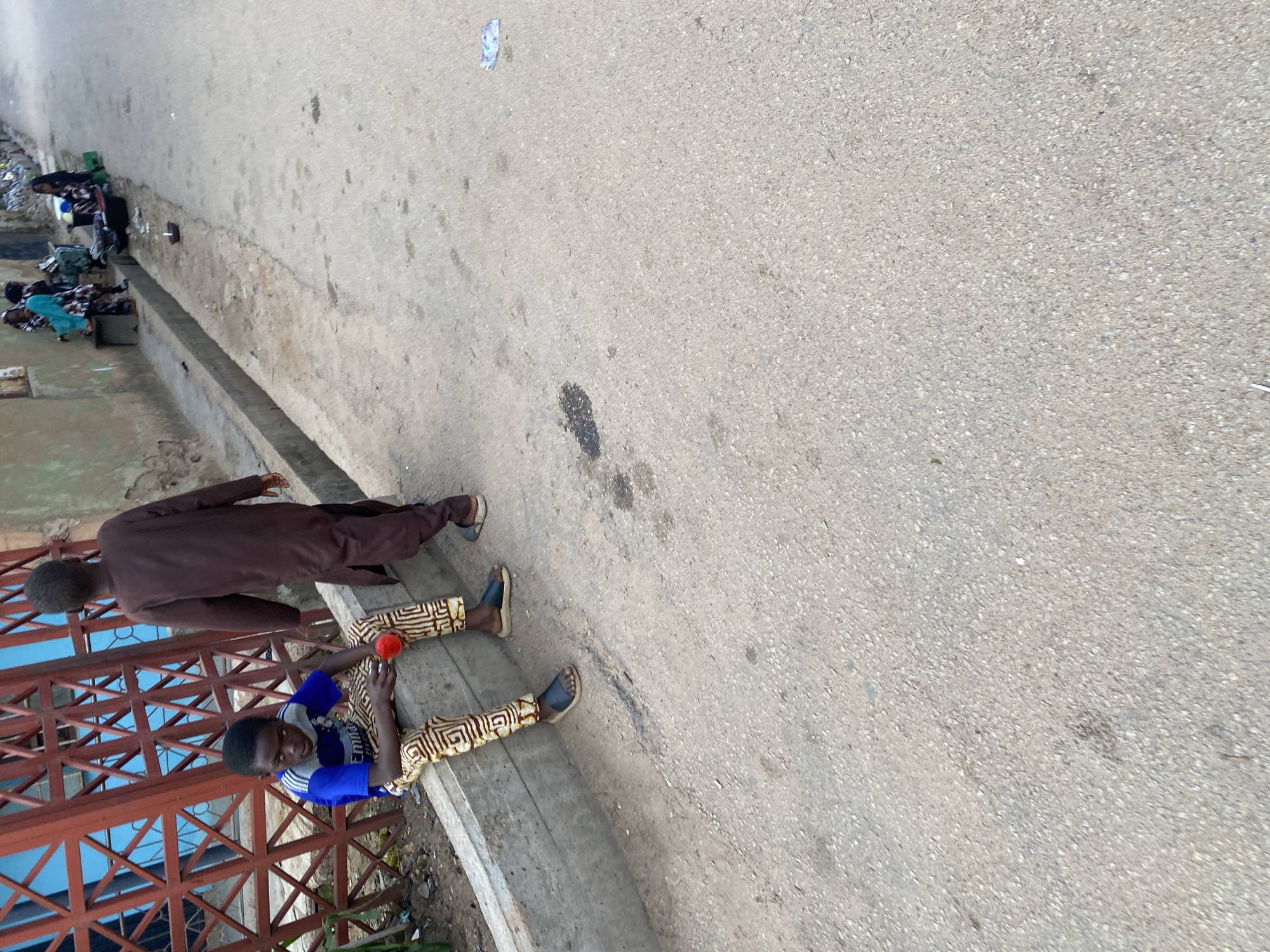 The image depicts a street scene in the city of Jos, Nigeria. In the foreground, a young boy in a blue shirt and patterned pants is sitting on a concrete curb, holding a red object, possibly a cup. To his right, another boy in a brown tunic walks away from the camera. Behind them, a group of women, some in colorful headscarves, are gathered near a wall, with various items laid out, suggesting a market or roadside vending activity. The environment consists of a paved road, a concrete drainage channel, and a building with a red metal fence in the background. The scene appears to be an ordinary day in a bustling urban area.