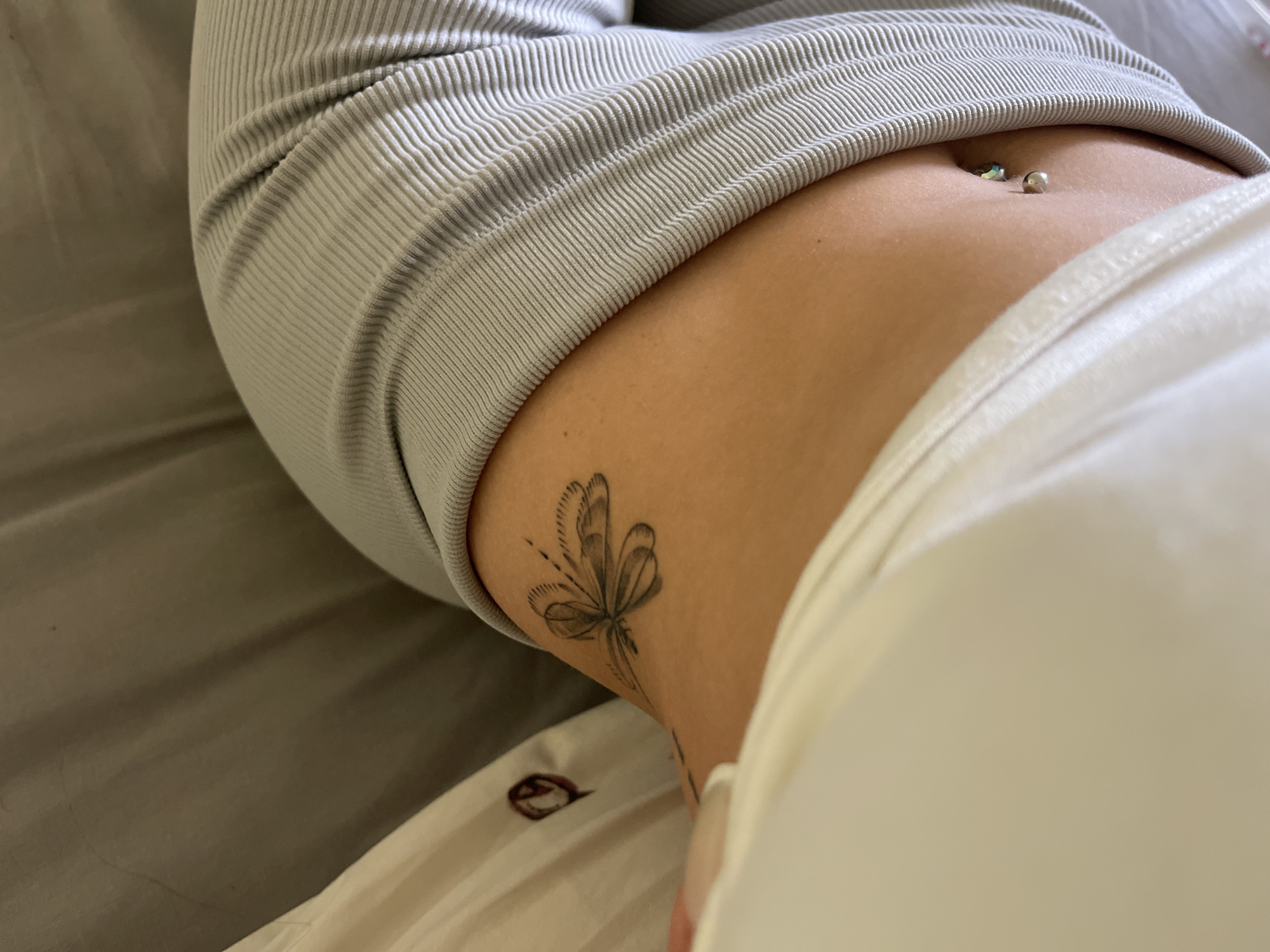 A close-up, high-angle shot shows a person's torso and side. The person is lying down, partially obscured by white fabric on the left and a ribbed grey fabric on the right. A black dragonfly tattoo is visible on the person's upper back or shoulder area, near the nape of the neck. A small belly button piercing with two charms is also visible. The background is a muted, textured grey surface. The scene is indoors, in an unknown location.