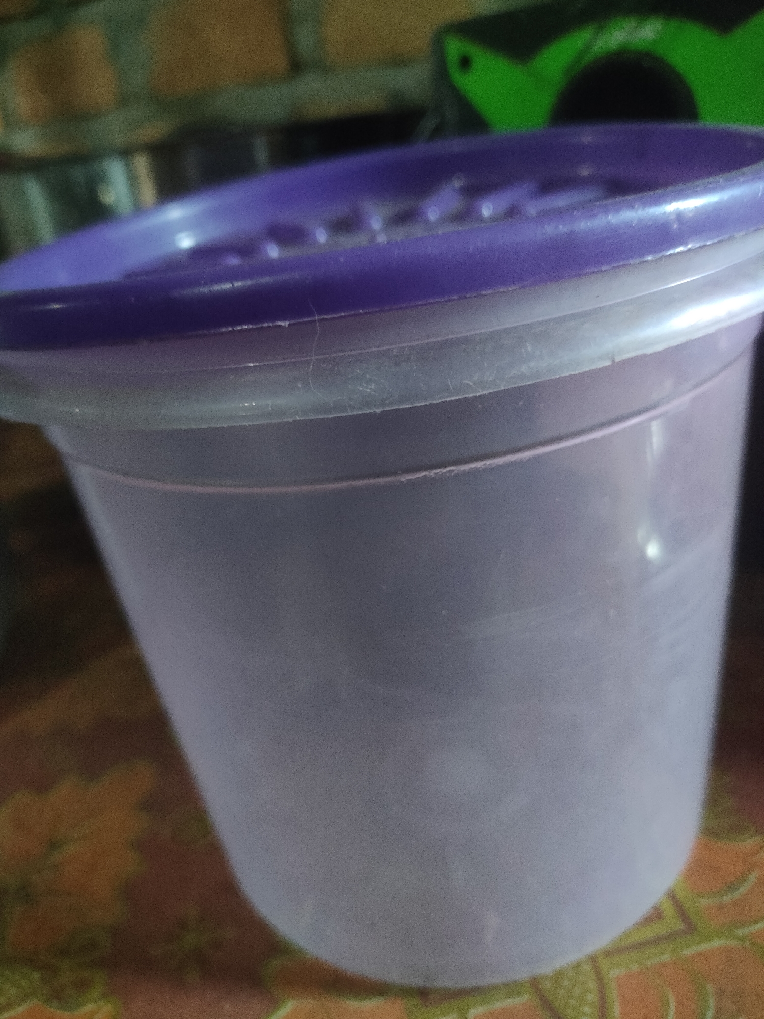 A close-up, slightly out-of-focus shot shows a purple plastic lid on a translucent purple plastic container. The container is cylindrical and appears to be about half full. The background is blurred and includes hints of a brick wall and a green object. The surface the container is resting on is textured and appears to be a table with an orange and green pattern. The image quality is poor with visible noise and lack of sharp focus. The location in the city of Padang Dalom, Indonesia, is not discernable from the image itself.