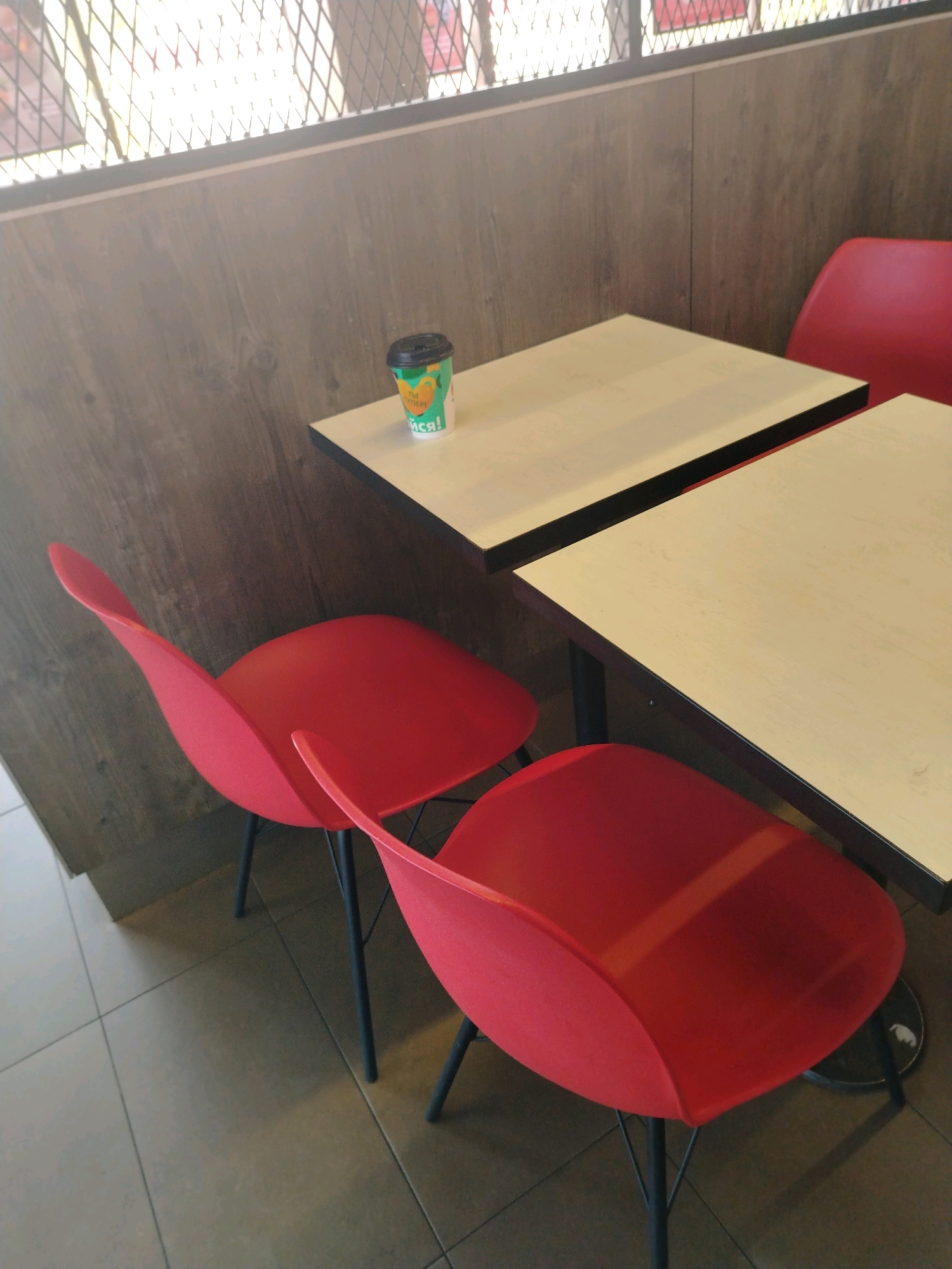 A paper coffee cup with a black lid sits on a light-colored table with a black edge. The cup has green and orange graphics and text that appears to be in Cyrillic script, possibly saying "Ты супер!" or "You are super!". Two red chairs with black legs are positioned around the table. The scene is indoors, with a wooden-paneled wall behind the table and chairs, and tiled flooring. A metal grate is visible in the background. The lighting suggests an overcast day or indoor lighting, with some glare on the grate. This image was taken in Yaroslavl, Russia.