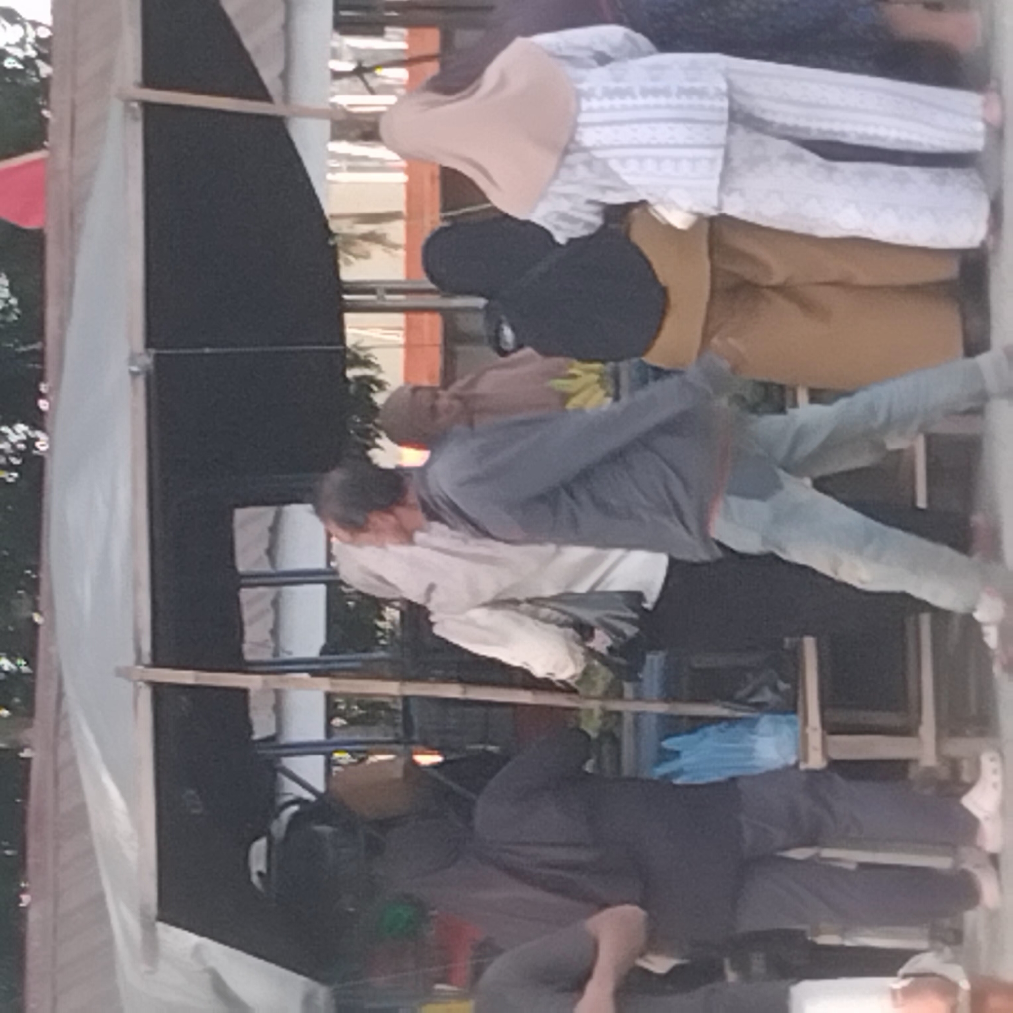 In Pelaihari, Indonesia, a group of people stand in front of a makeshift market stall. A woman in a light-colored hijab and white patterned clothing stands facing away from the camera. To her left, a person in a dark hijab and brown pants is also visible. In the foreground, a person in a gray jacket and light blue jeans is walking, with their back to the camera. To the far left, a person wearing a dark gray shirt and dark pants is partially visible. A wooden stall structure supports a tarp overhead, creating a shaded area. Various objects, including blue plastic bags, are visible on or near the stall. The overall scene appears to be an outdoor market environment.
