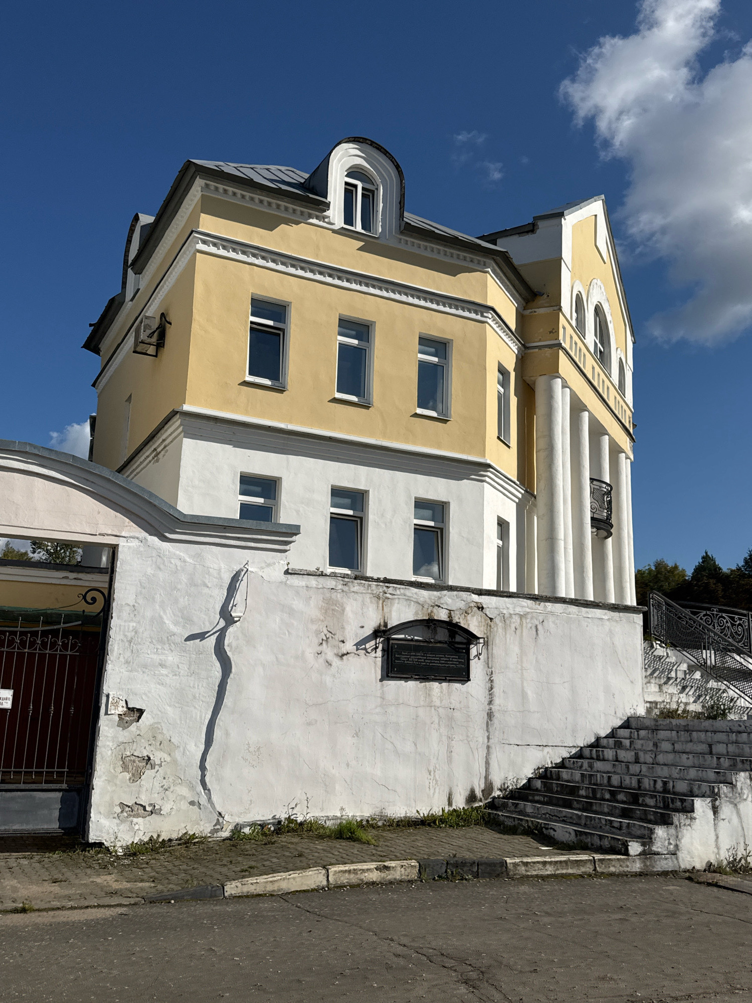 The image depicts a building with a yellow upper story and a white lower story, situated in Torzhok, Russia. The building features arched windows on the upper floors and a series of white columns supporting a portico on the right side. In the foreground, there is a white wall with peeling paint, a metal gate on the left, and a set of stone stairs leading up the right side. A plaque is affixed to the wall below the stairs. The sky is blue with some white clouds, and the ground is paved with asphalt and interlocking bricks. The lighting suggests it is daytime, likely morning or afternoon, given the distinct shadows.