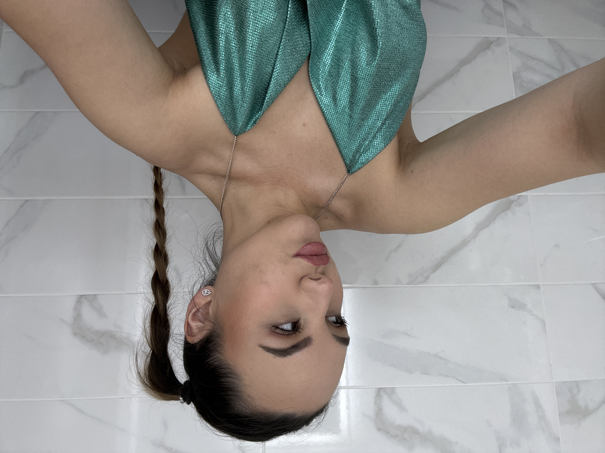 The image depicts a woman with fair skin and brown hair styled in a braid and low ponytail. She is wearing a teal, shimmery top. The background shows a white tiled wall or floor with grey veining, resembling marble. There is no evidence of this photo being taken in Sofia, Bulgaria.