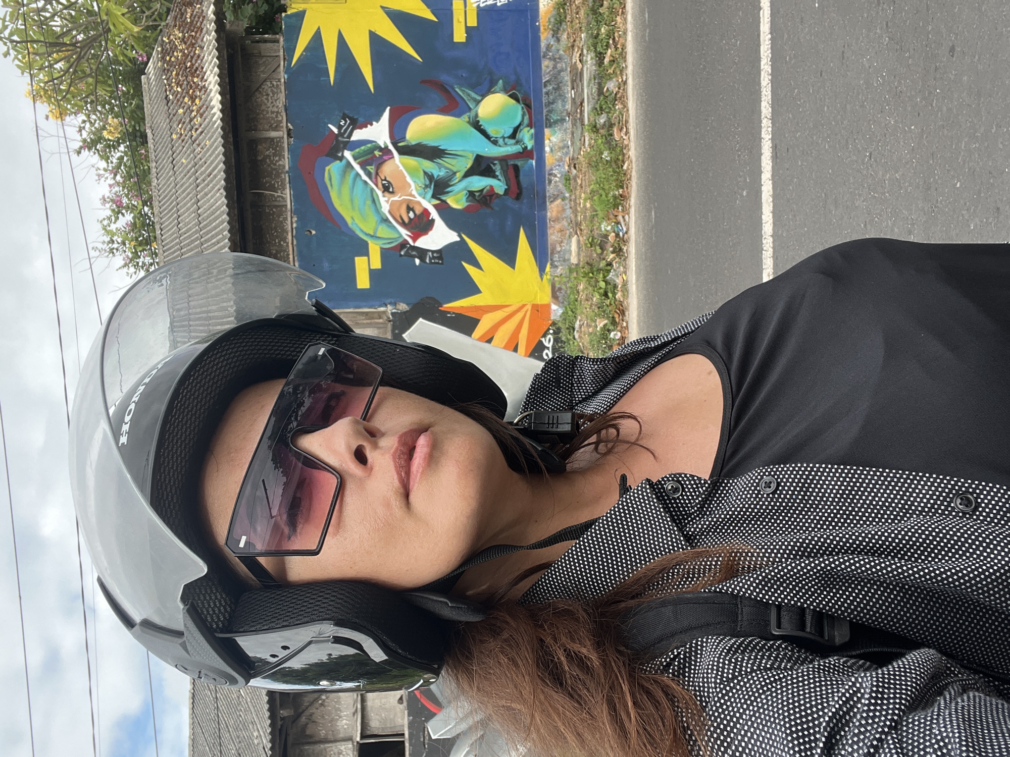 A woman wearing a black helmet, large sunglasses with pink lenses, and a black and white polka dot shirt over a black tank top is captured in a selfie. She is on a road with a graffiti-covered wall in the background. The graffiti depicts a stylized figure in shades of blue and green, with yellow bursts. The scene appears to be outdoors, with a cloudy sky, a roofline with grey tiles, and some greenery. A portion of the road with a white line is visible in the foreground, suggesting Kuta Selatan, Indonesia.