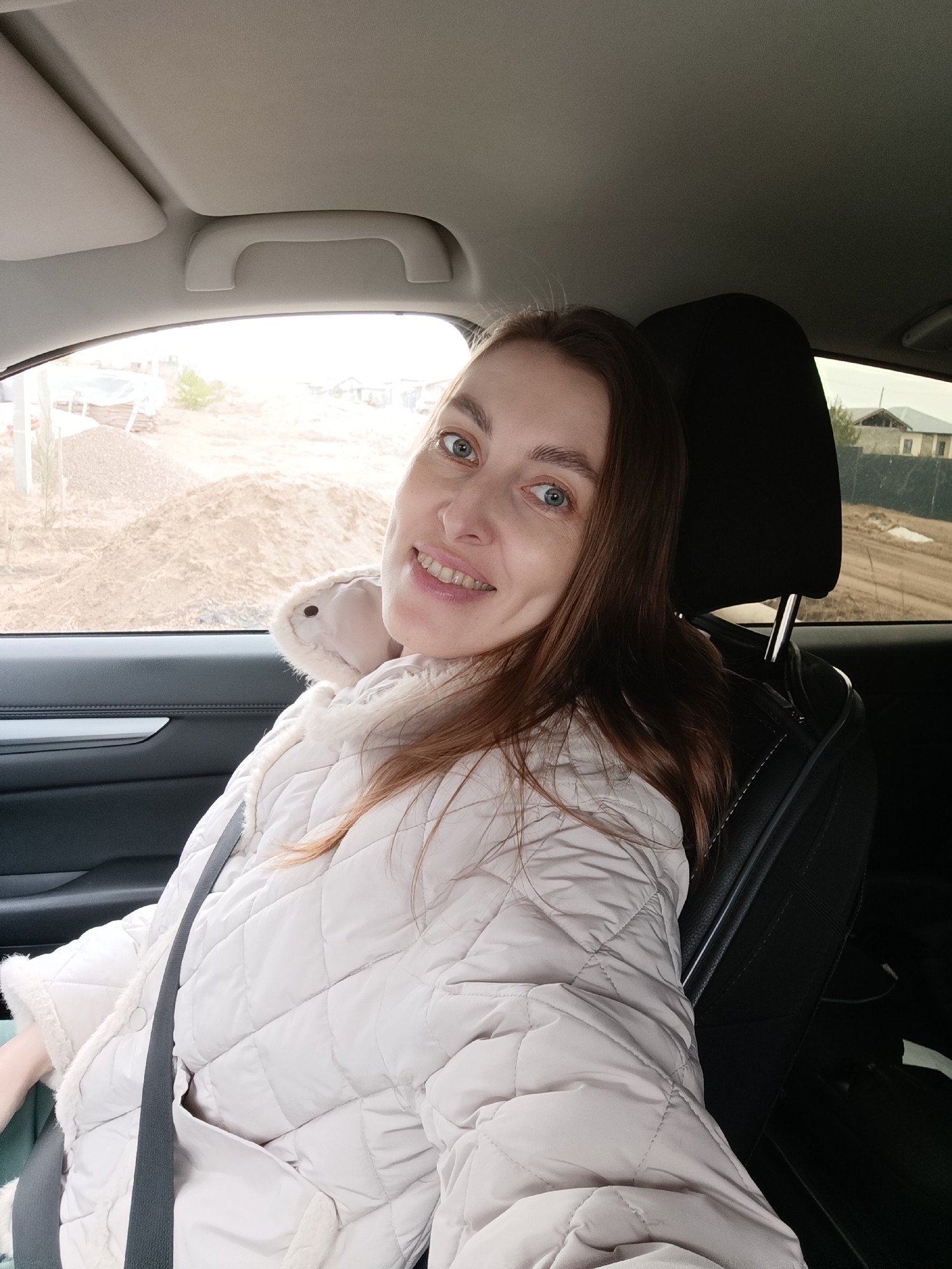 A woman is sitting in the driver's seat of a car, wearing a white quilted coat with fur trim and a seatbelt. She is smiling and looking at the camera. Outside the car window, there's construction or landscaping activity with piles of dirt. Beyond that are houses, suggesting a residential area. The scene is located in what looks like Chaikovsky, Russia.