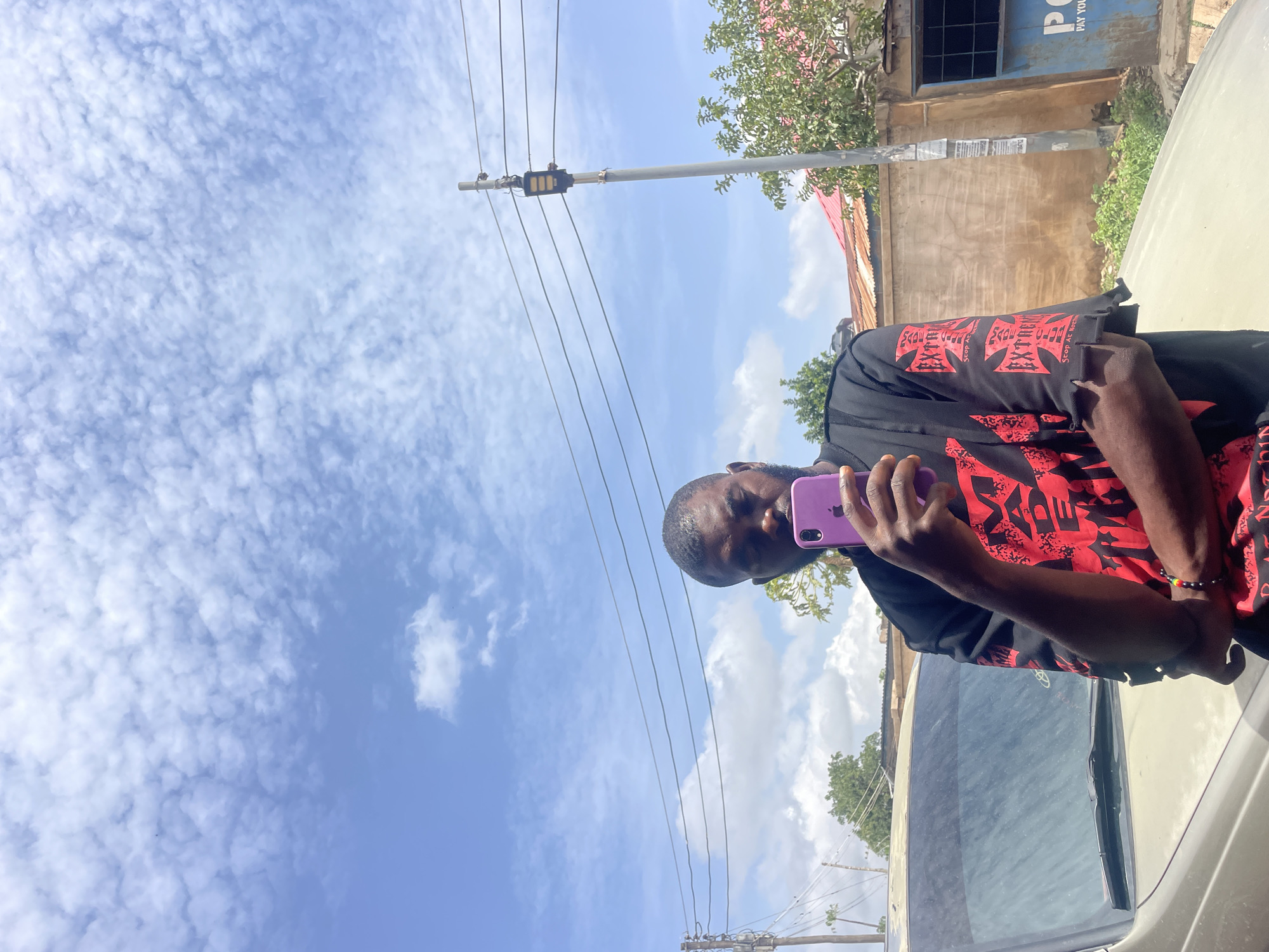 A medium shot shows a man holding a purple smartphone and looking toward the camera. He is wearing a black t-shirt with a red design, and an unidentified vehicle’s hood and windshield are visible to his right. The background includes power lines, a utility pole, buildings, and trees. The sky is predominantly blue with scattered clouds. Based on the "PC PAY YOUR" sign in the background, this is in the city of Jos, Nigeria.
