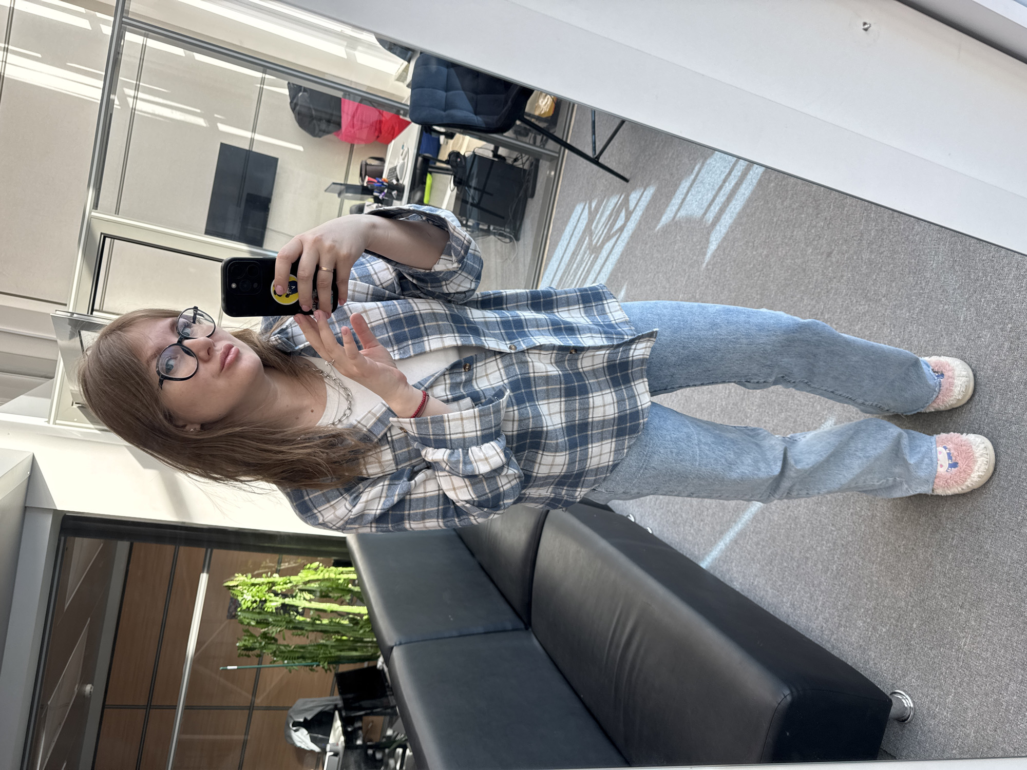 A female subject is depicted taking a mirror selfie within an indoor commercial or office environment. The subject has light brown hair and wears black-framed glasses, a white undershirt, an unbuttoned blue, white, and beige plaid long-sleeved shirt, light blue jeans, and pink and white fuzzy slippers with a cartoon motif. She wears a silver chain necklace, a ring on her right hand, and a red bracelet on her left wrist. She holds a black smartphone, featuring a yellow circular smiley-face sticker, in her left hand, directed at a large vertical mirror that reflects her image. The immediate environment includes a grey carpeted floor.
The background, visible through the mirror's reflection, reveals an office setting. To the subject's left is a black leather two-seater sofa with chrome legs. Beyond the sofa, a tall green columnar plant stands in a dark pot. The wider background consists of glass partitions, wooden wall panels, and open office areas. Reflected elements include a white desk with a computer monitor, a dark office chair, and a bright pink object (possibly a bag or garment). Natural light streams into the space, casting distinct diagonal shadow patterns from window frames onto the carpet and walls. The specific location within Perm, Russia, cannot be determined from the visual information alone.