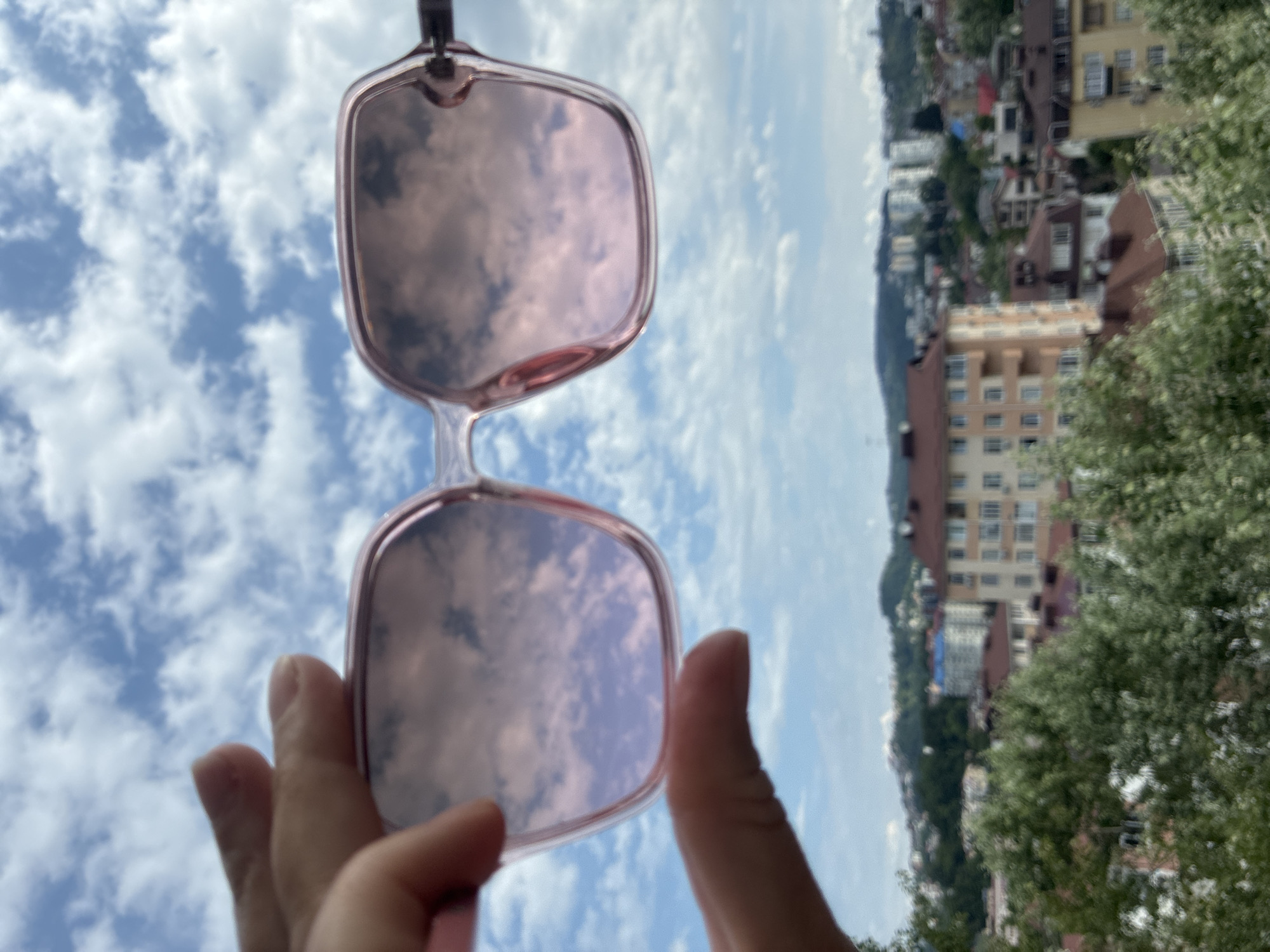 A hand holds up a pair of pink-tinted sunglasses against a blue sky with white clouds. The lenses of the sunglasses reflect the sky and clouds, creating a mirror image. Below the sky and sunglasses, a blurred cityscape with buildings, trees, and hills is visible. The buildings are a mix of residential and commercial structures with red and beige roofs. The overall scene suggests a view from a high vantage point in Sochi, Russia.