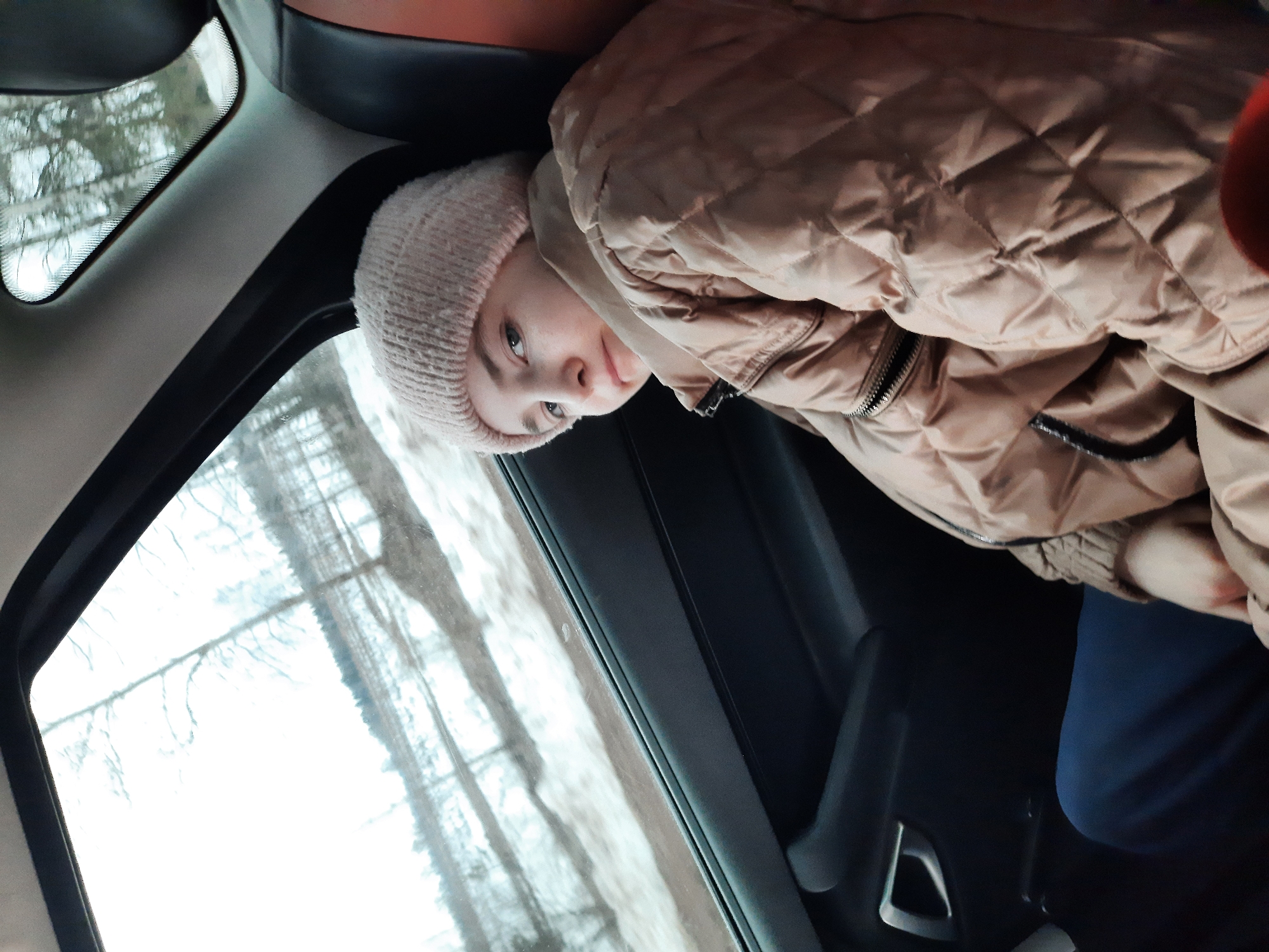 A young person wearing a pink knit hat and a tan quilted jacket is seated in a car, looking towards the viewer. The car window shows a blurred view of trees, suggesting an outdoor setting, possibly in a rural or suburban area. Given the city name, the image was likely captured in or near Архангельское, Russia. The interior of the car is dark, highlighting the individual near the window. The person's expression is neutral. The lighting is natural, coming from the window. The person appears to be a passenger in the car.