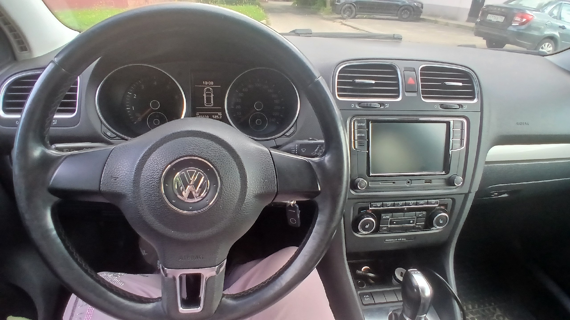 The image shows the interior of a Volkswagen car, likely a Golf, viewed from the driver's seat. A key is inserted in the ignition. The dashboard displays the time as 10:00 and the odometer reads 285030.585.2. The steering wheel has the Volkswagen logo. The car is parked on a street, with other cars visible outside. Based on the license plate of the Lada Granta in the background, which reads "303-777-190 RUS", this image was likely taken in the city of Mozhaisk, Russia. The environment appears to be an urban setting during the daytime, with buildings and greenery visible in the background.