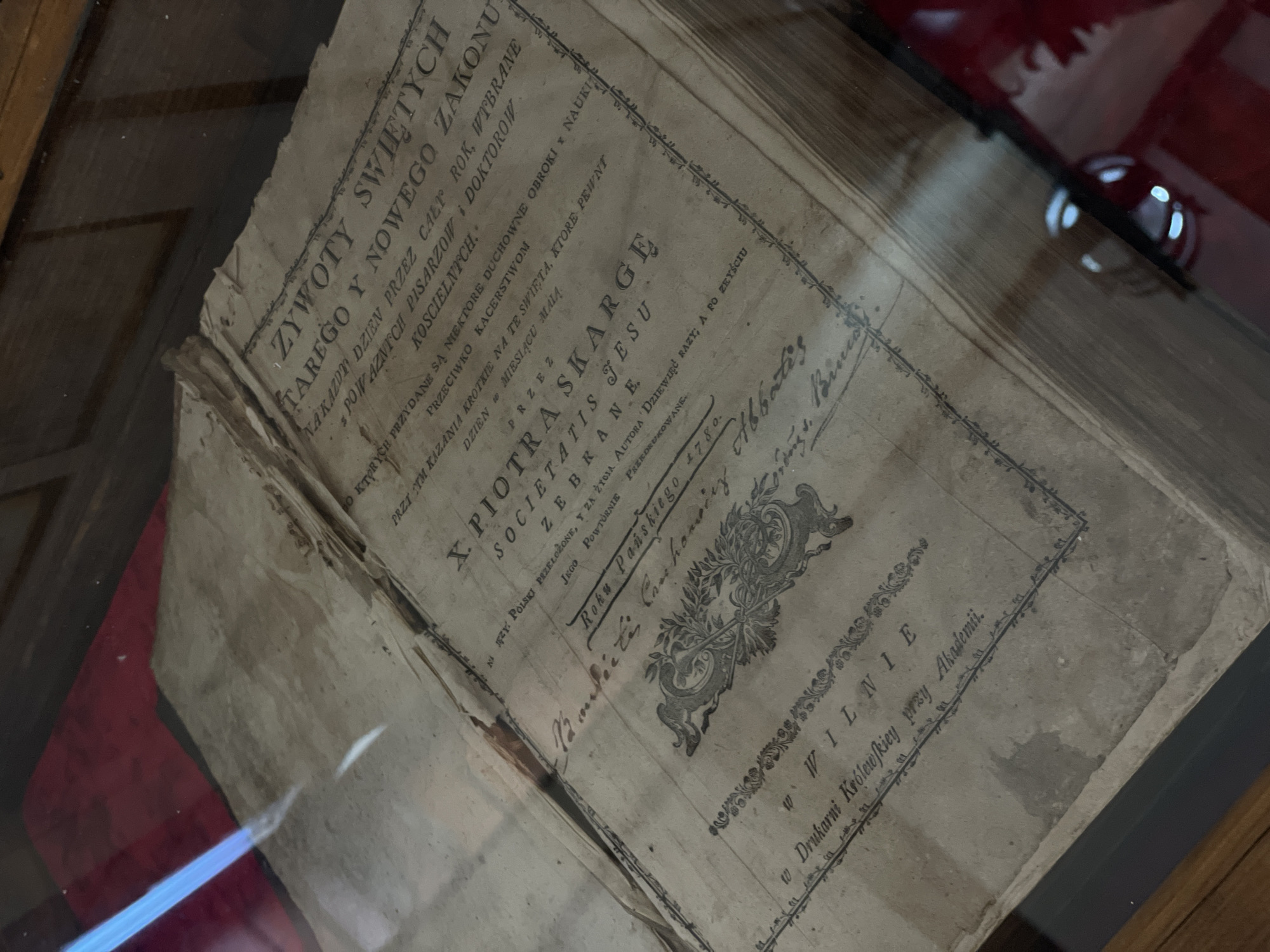 The image shows an open, antique book with yellowed pages and visible wear. The title page, written in an old script, is the main focus. The text appears to be in Polish, with phrases like "ŻYWOTY ŚWIĘTYCH" and "SKARGE JESTU". There is a decorative border around the text. Below the title, there are inscriptions and what appear to be handwritten signatures. The book is placed on a surface, possibly a display case, with reflections on the glass. The environment suggests an indoor setting, possibly a library or museum. The specific location in Літвенскі сельскі Савет, Belarus cannot be determined from the image.