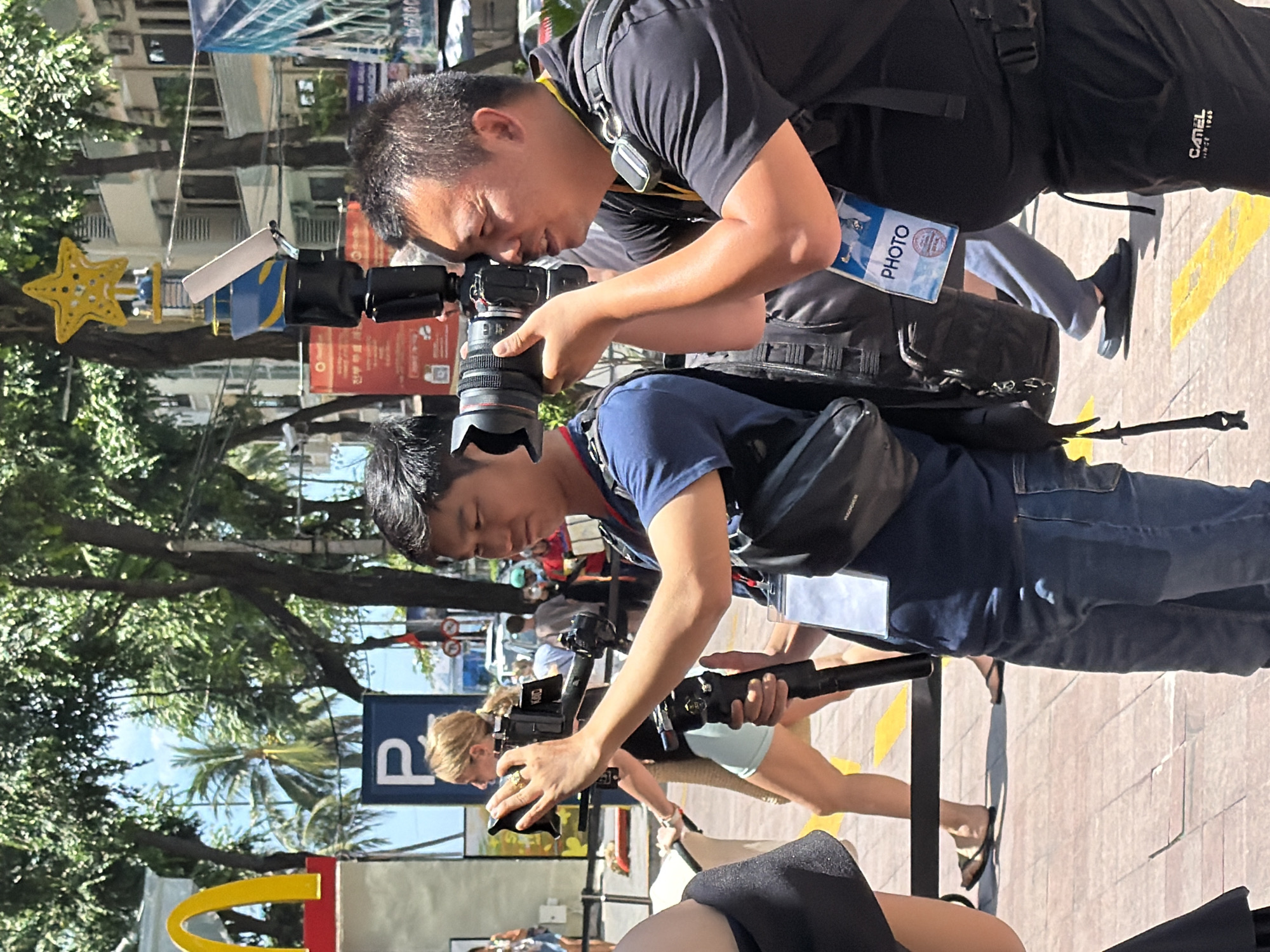 The image depicts an outdoor scene in Nha Trang, Vietnam, likely on a street or public walkway. In the foreground, two men are actively engaged in photography. The man on the right, wearing a black t-shirt and black pants, is holding a professional camera with a large lens and a flash unit. He is looking through the viewfinder and appears to be focusing on something outside the frame. He is wearing a lanyard with a "PHOTO" badge around his neck. The man to his left, wearing a blue t-shirt and blue jeans, is also holding a camera on a rig, aiming it forward. His focus is also directed away from the camera.
Behind the photographers, a woman with blonde hair tied in a ponytail is visible, wearing a black top and light blue shorts. She is walking away from the viewer, holding what appears to be a straw bag. Further in the background, other people are present, some walking and some standing, suggesting a public area with some activity. Palm trees and leafy green trees are prominent in the background, indicating a tropical or subtropical environment. A portion of a McDonald's restaurant signage, featuring the iconic golden arches, is visible on the left side of the image, suggesting the location is near a commercial area. Buildings, possibly hotels or shops, can be seen in the distance, along with a blue sign with a white "P" on it. The ground is paved with interlocking bricks and has yellow markings. The overall impression is of a bright, sunny day.