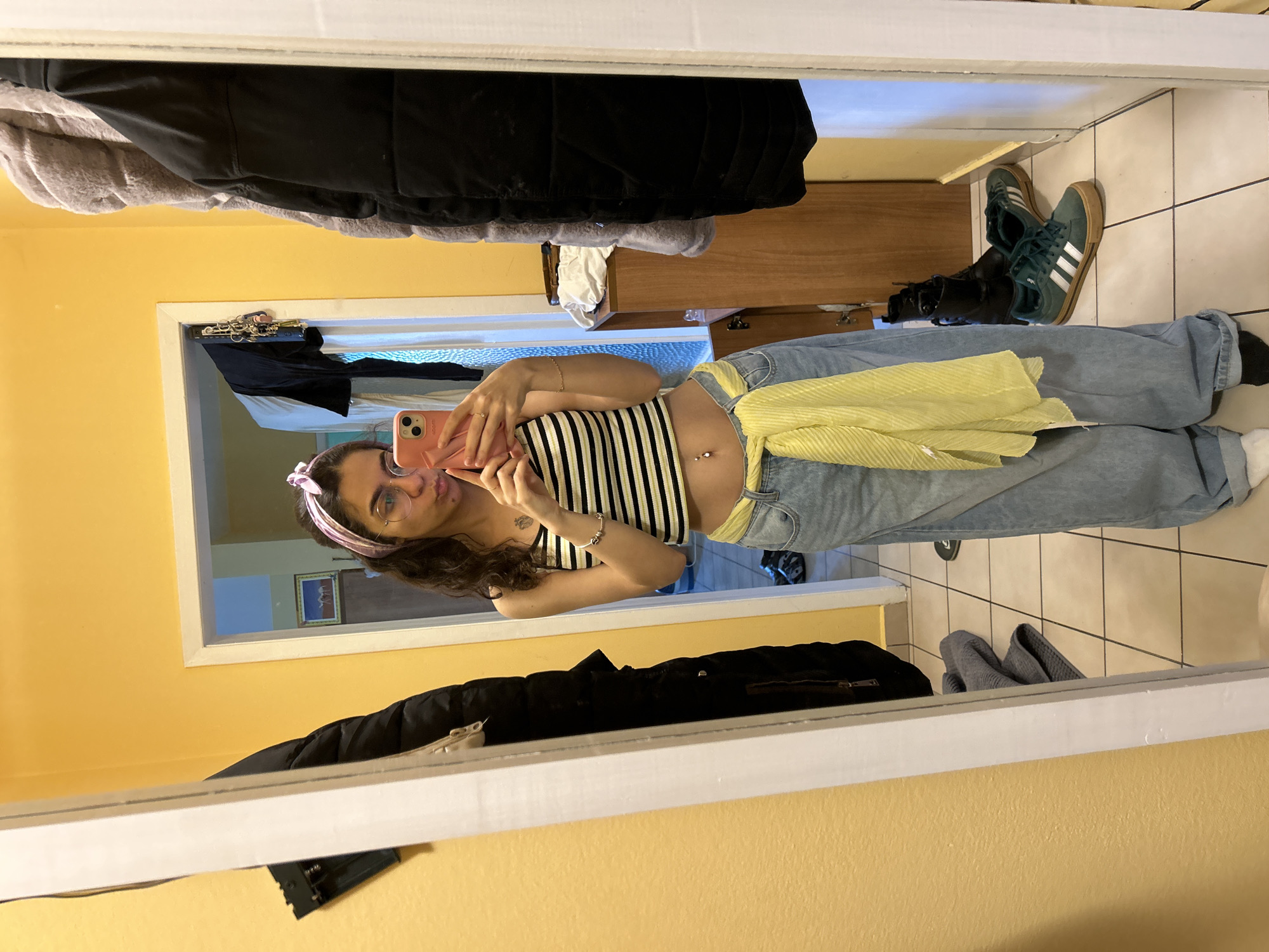 A young woman is taking a mirror selfie indoors. She is wearing a striped crop top, loose blue jeans with a yellow garment tied around the waist, and a pink headband. She is holding a pink mobile phone. The mirror reflects a hallway with a partially open doorway leading to another room. In the hallway, there is a cabinet or dresser, shoes on the floor, and coats hanging. The walls are yellow. The location cannot be determined as being in Sofia, Bulgaria without additional information.