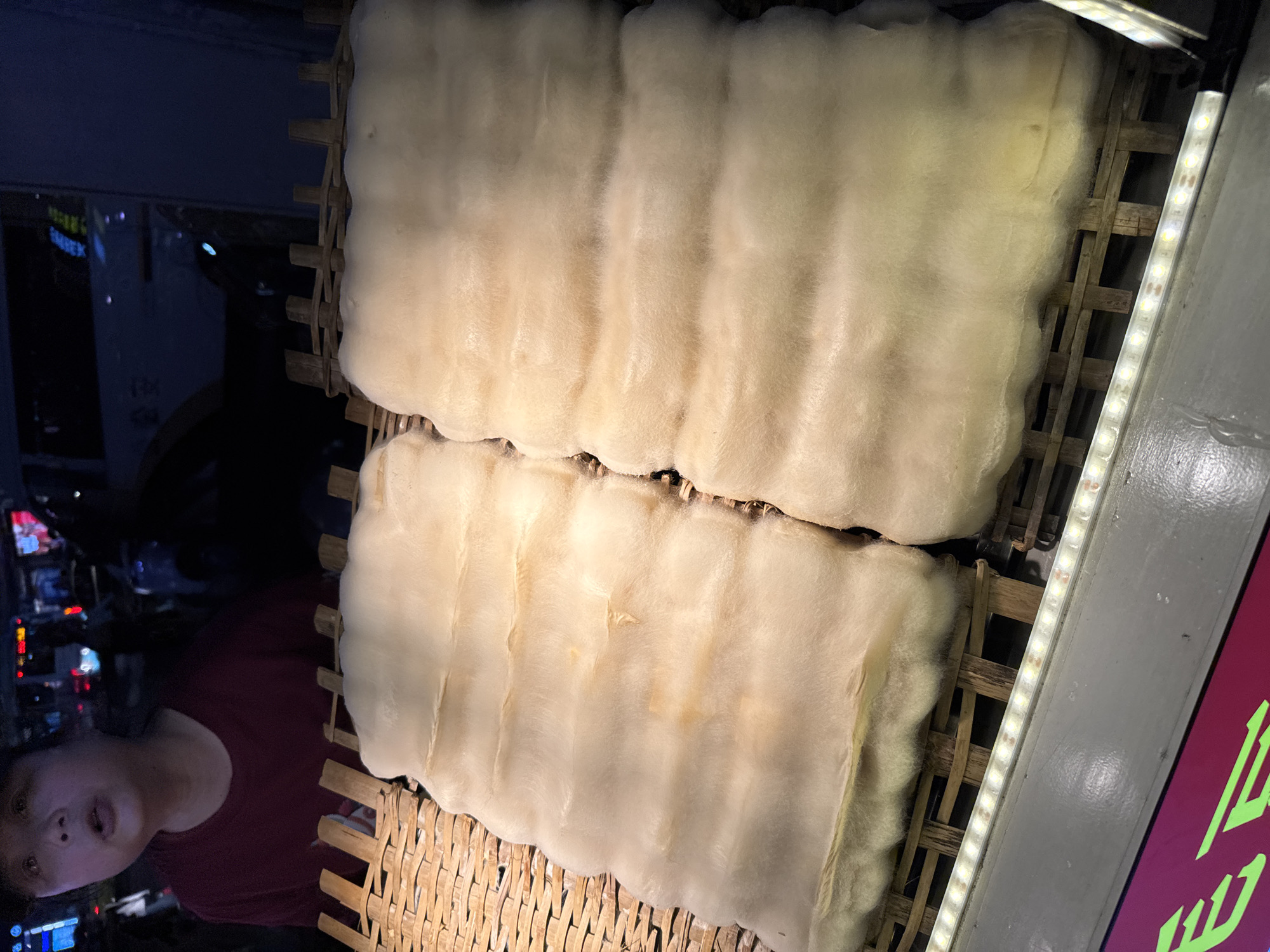 The image shows two large rectangular blocks of a white, fluffy substance, possibly cotton candy or a similar confection, displayed on a woven bamboo or wooden frame. The scene is lit by a strip of LED lights, which are positioned on the right edge of the image and cast a bright glow. A portion of a person's face, specifically their chin, cheek, and the side of their neck and upper torso, are visible in the lower-left corner, appearing to be behind the display. A red shirt or garment is also visible. The background reveals an out-of-focus street scene with vehicles, lights, and possibly buildings, suggesting an urban environment. The presence of Chinese characters (a red sign) suggests the location is likely in a Chinese-speaking area, which aligns with the Yuzhong District, China specification.