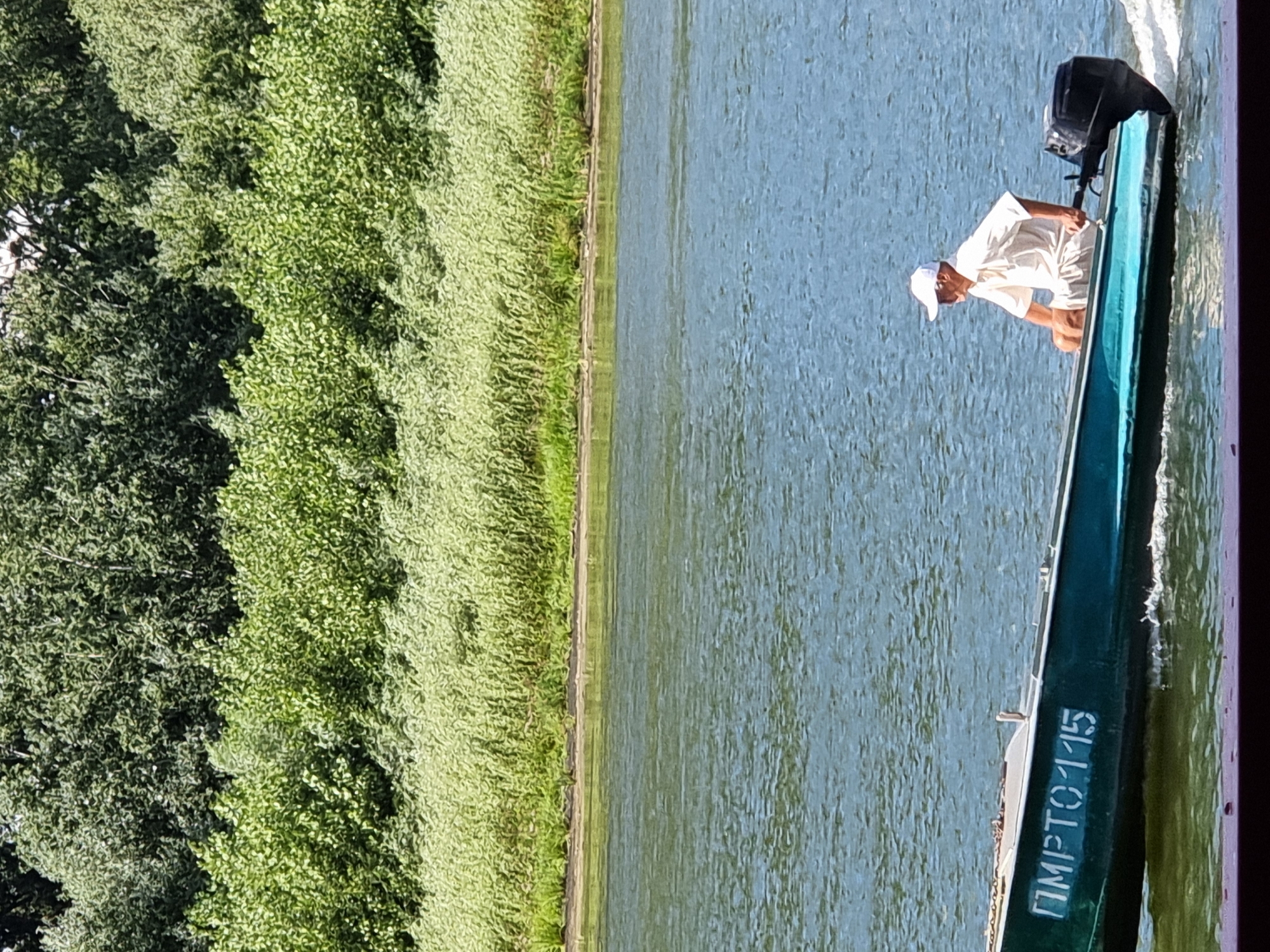 In the city of Kitskany, Moldova, a teal-colored boat with the identification number "ПМРТО 115" inscribed in white lettering on its side travels across a body of water. A man wearing a white cap and a light-colored shirt is seated in the boat, which is propelled by a black outboard motor. The background features a dense line of green trees and vegetation along the bank of the river. The water's surface is rippled, indicating movement.