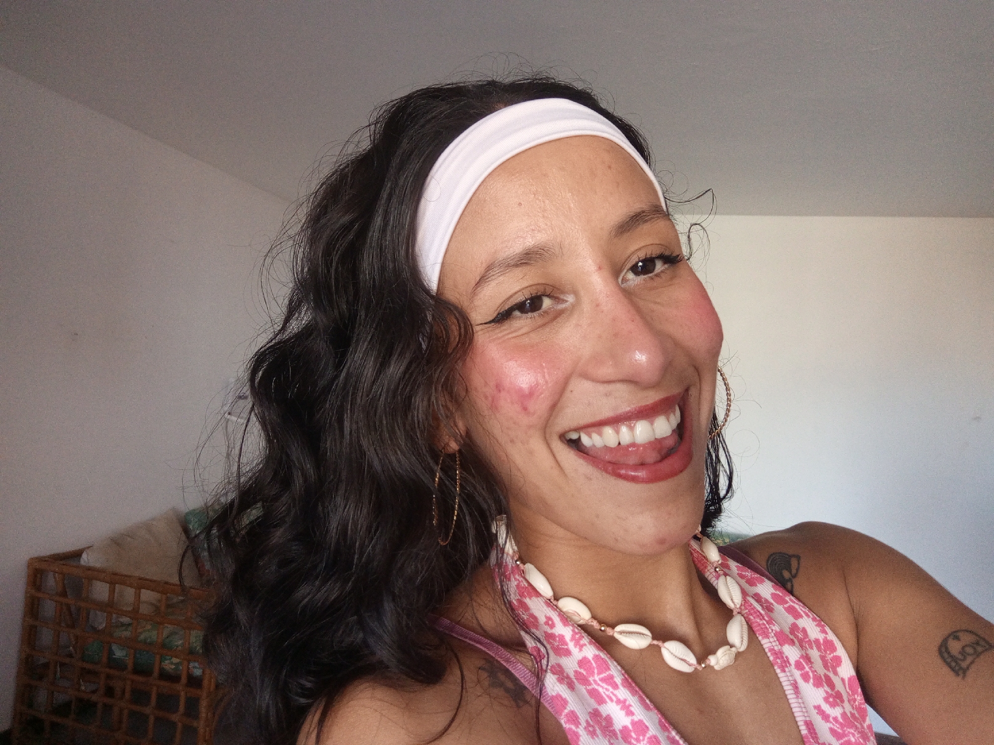 A woman is pictured indoors with a white wall behind her. She has long dark hair, a white headband, and is wearing a pink halter top with white flowers, and a shell necklace. Gold hoop earrings adorn her ears. She is smiling, showing her teeth. A woven basket is partially visible in the lower left corner. Tattoos are visible on her right arm. The scene does not provide enough information to determine if the location is in Porlamar, Venezuela.
