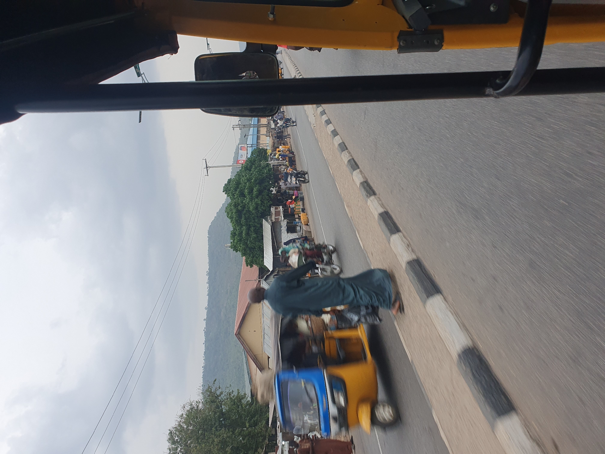 The image depicts a street scene in Lokoja, Nigeria, viewed from inside a moving vehicle. A yellow and blue "keke" (auto rickshaw) is visible in the foreground, being driven along a paved road with black and white striped curbs. A man wearing a dark green robe appears to be a passenger, with a blur indicating movement. In the background, there are other vehicles, people, and roadside stalls, suggesting a busy commercial area. A mountain is visible in the distance under a cloudy sky. The overall environment appears to be a typical developing urban landscape. Electrical power lines are visible in the sky.